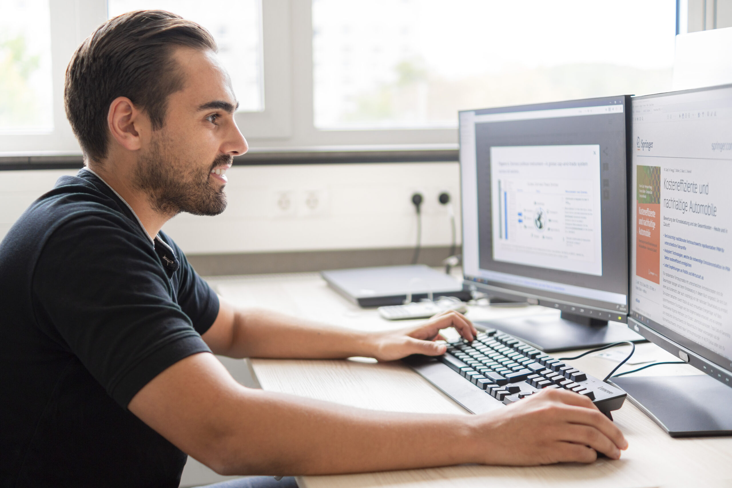 Ein Mann sitzt an einem Schreibtisch und konzentriert sich auf zwei Computermonitore, auf denen Grafiken und Texte angezeigt werden. Durch das Fenster hinter ihm fällt Tageslicht in seinen Arbeitsbereich. Als Absolvent der Hochschule Coburg wendet er sein erlerntes Fachwissen an, um Daten mithilfe von Tastatur und Maus effizient zu analysieren.