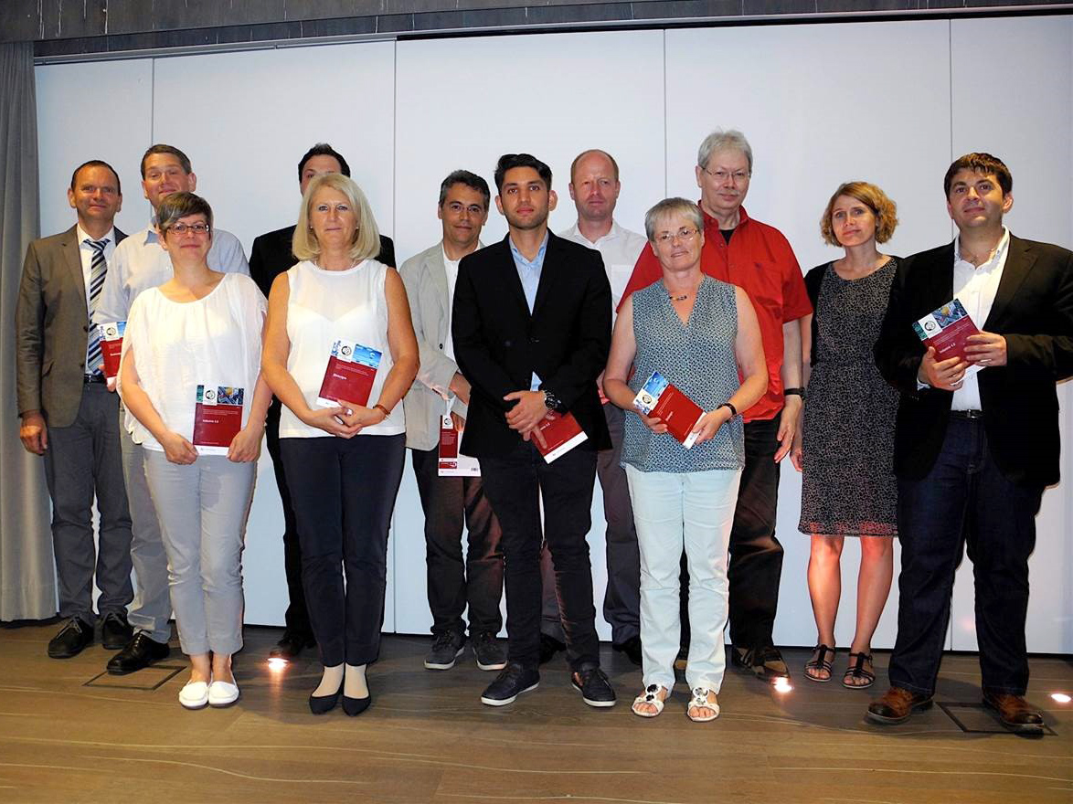 Eine Gruppe von zwölf Personen, darunter Männer und Frauen, steht in zwei Reihen. Einige tragen halbformelle Kleidung und halten rote Notizbücher in den Händen. Der Hintergrund ist eine schlichte weiße Wand, die auf ein akademisches Umfeld wie die Hochschule Coburg hindeutet.