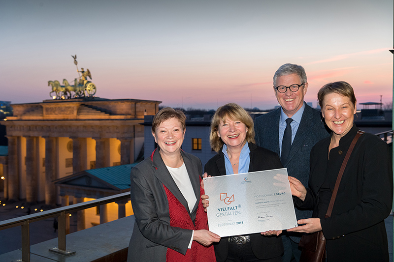 Vier Menschen stehen in der Abenddämmerung auf einer Terrasse, mit dem ikonischen Brandenburger Tor im Hintergrund, strahlend vor Stolz. Unter klarem Himmel halten sie eine Urkunde der Hochschule Coburg in den Händen und würdigen gemeinsam diese denkwürdige Leistung.