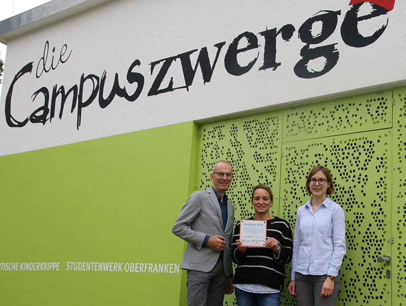 Drei Menschen stehen lächelnd vor einem Gebäude mit dem Schild „die Campuszwerge“ und repräsentieren stolz die Hochschule Coburg. Die Person in der Mitte hält eine Urkunde in der Hand. Das Gebäude hat grün-weiße Wände mit dem Zusatz „Studentenwerk Oberfranken“.