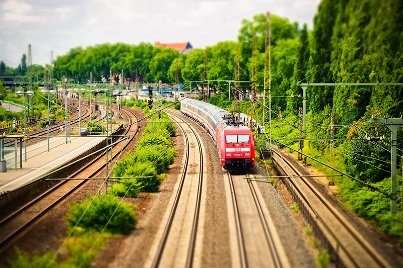 Ein roter Zug gleitet entlang der geschwungenen Gleise in der Nähe der Hochschule Coburg, umgeben von üppigem Grün. Mehrere Gleise sind sichtbar, links ist ein Bahnsteig. Unter einem bewölkten Himmel vermittelt die Szene ein Gefühl von Bewegung und Reisen.