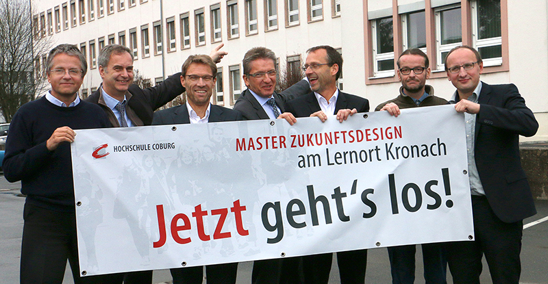 Sieben Männer in Anzügen und Freizeitkleidung stehen vor einem Gebäude und halten ein Banner mit der Aufschrift „Master Zukunftsdesign am Lernort Kronach. Jetzt geht’s los!“. Sie wirken gut gelaunt und repräsentieren stolz die Hochschule Coburg, als sie für das Foto posieren.