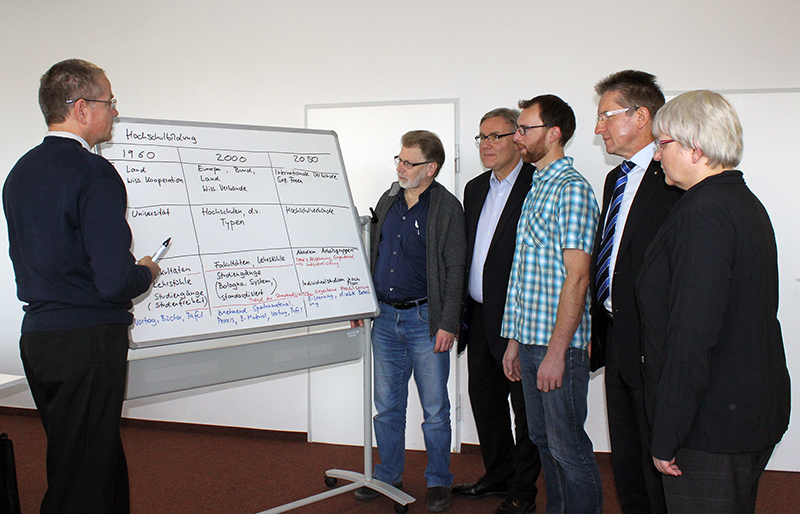 Eine Gruppe von sechs Erwachsenen steht an der Hochschule Coburg um ein Whiteboard. Eine Person schreibt etwas auf die Tafel, während die anderen zuschauen und diskutieren. Auf der Tafel finden sich zahlreiche handschriftliche Notizen und Diagramme, die auf ein produktives Meeting oder eine Präsentation in dem gut beleuchteten Raum schließen lassen.