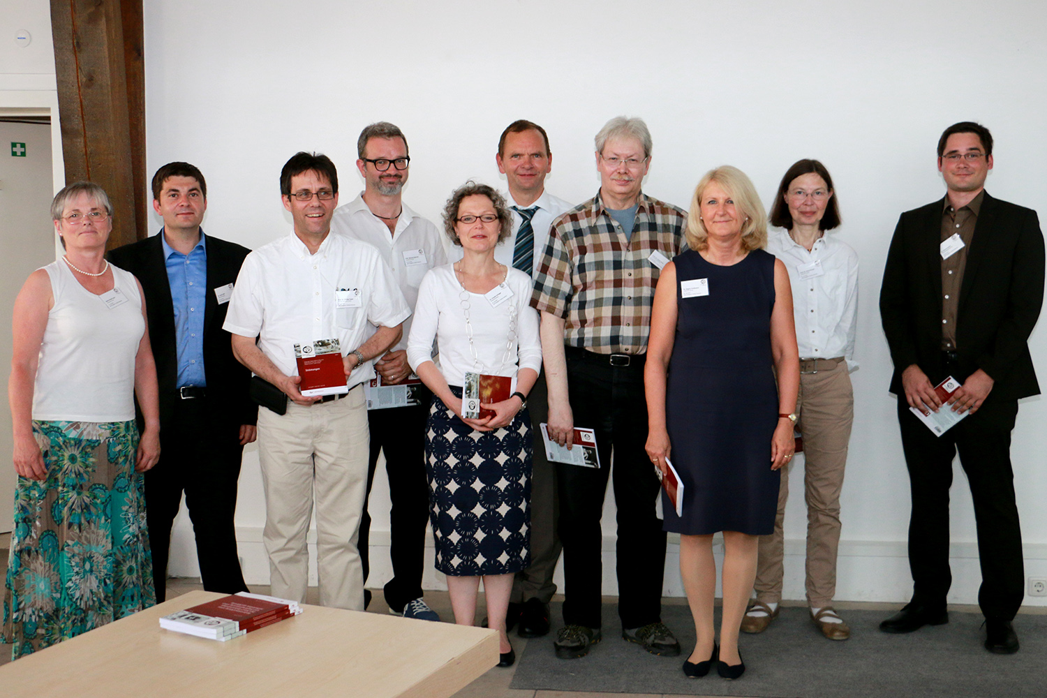 Eine Gruppe von zehn Personen steht in einem Gebäude der Hochschule Coburg und lächelt in die Kamera. Einige halten Bücher oder Broschüren in den Händen. Sie sind leger gekleidet, eine Mischung aus Männern und Frauen in verschiedenen Hemden, Hosen und Röcken, vor einer schlichten weißen Wand.