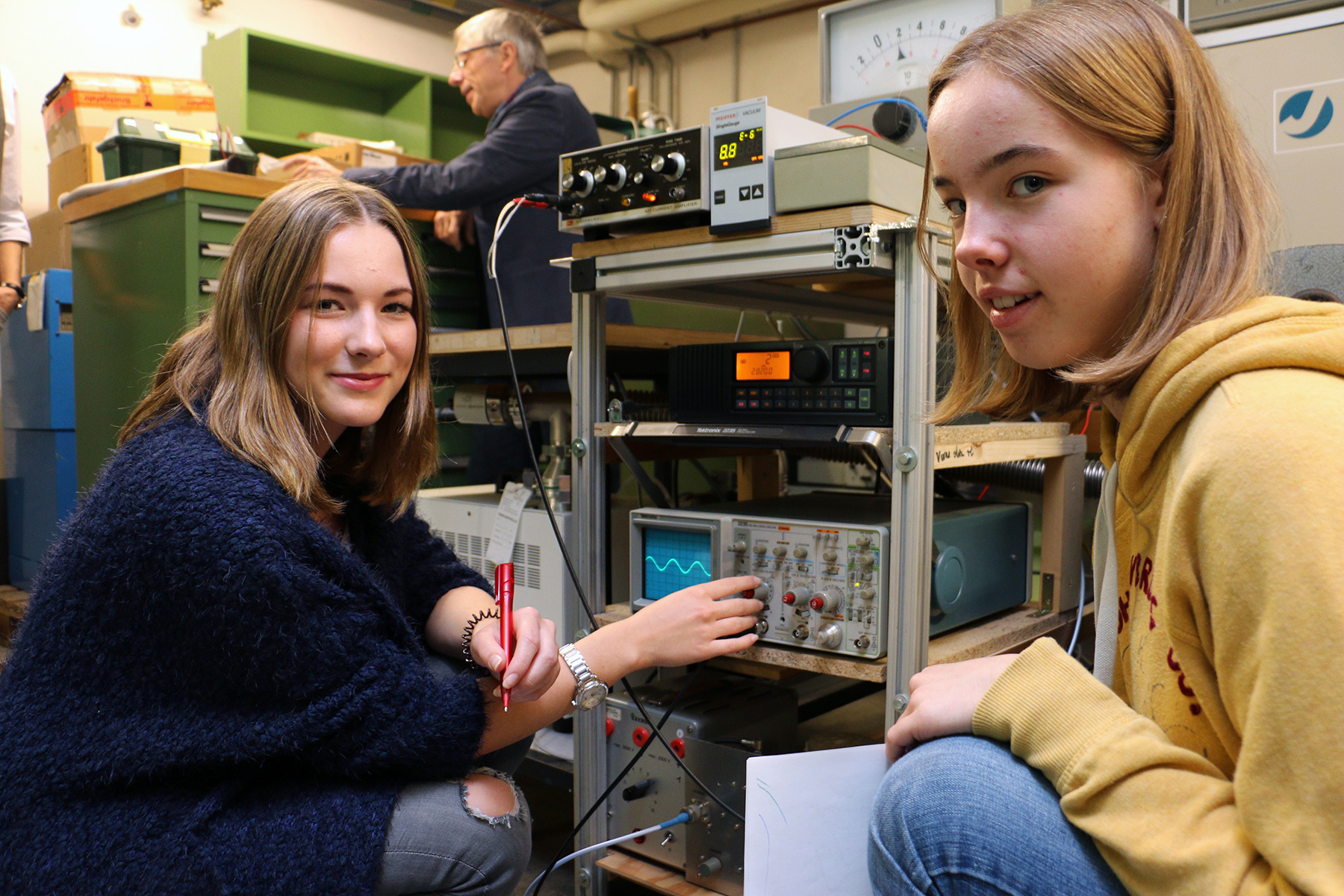 Zwei junge Frauen an der Hochschule Coburg sind in ihre Laborarbeit vertieft. Eine justiert ein Gerät, während die andere ein Blatt Papier hält. Im Hintergrund steht ein Mann an einem Regal, umgeben von verschiedenen elektronischen Geräten und Messinstrumenten, was die geschäftige akademische Atmosphäre verkörpert.