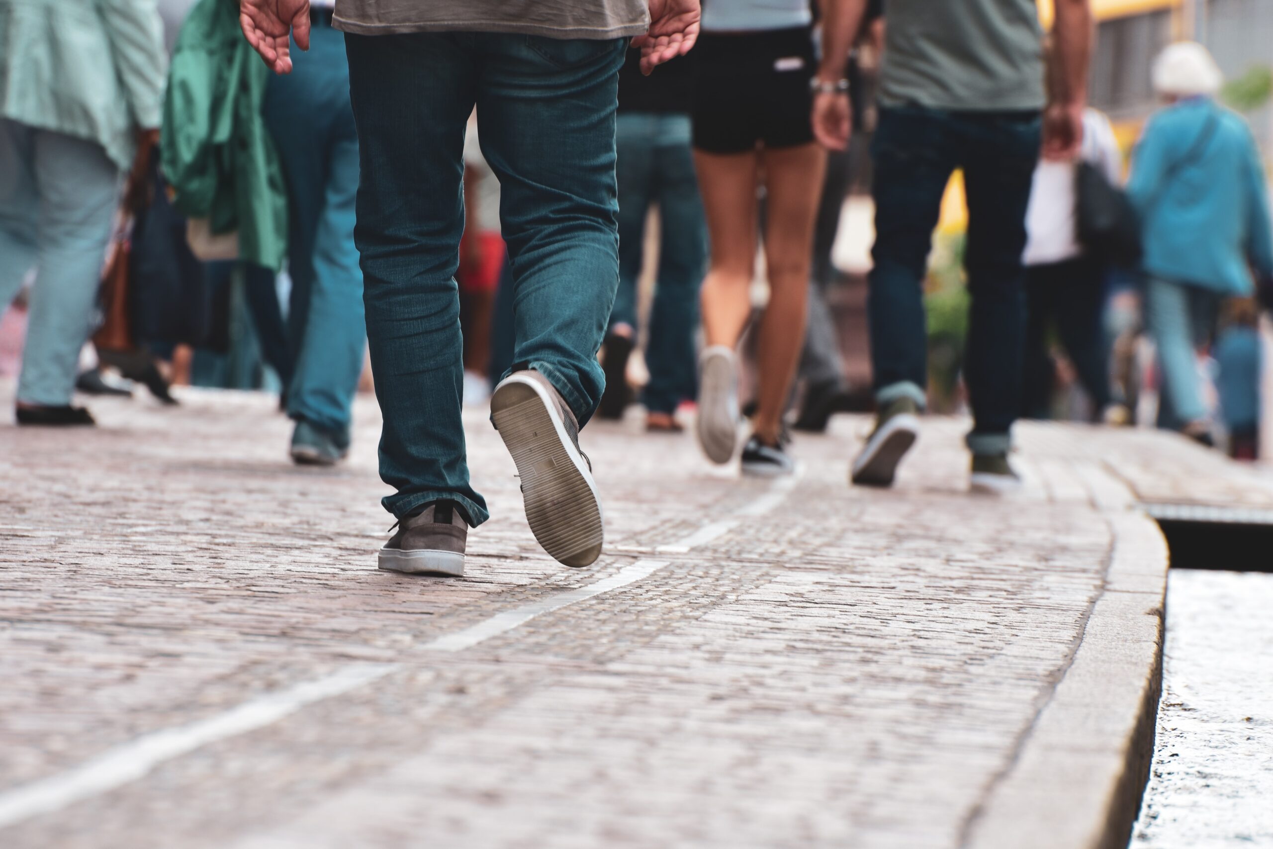 Eine Gruppe von Menschen, möglicherweise Studierende der Hochschule Coburg, läuft zügig über eine Kopfsteinpflasterstraße. Der Fokus liegt auf Beinen und Füßen in legerer Kleidung und Turnschuhen, die die dynamische Atmosphäre einer urbanen Umgebung einfangen.