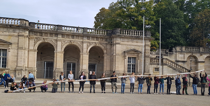 Eine Gruppe von Menschen steht in einer Reihe und hält ein außergewöhnlich langes Baguette vor der historischen Hochschule Coburg mit ihren majestätischen Bögen und Säulen. Üppige Bäume bilden eine malerische Kulisse.