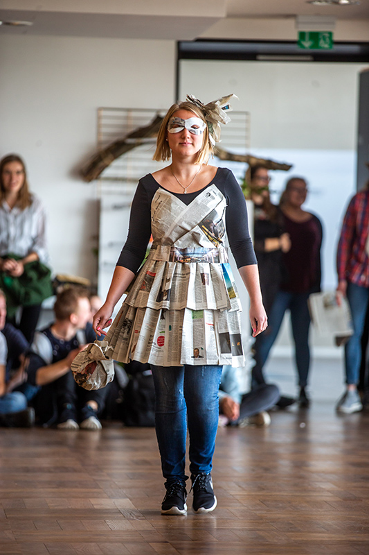 Eine Frau schreitet selbstbewusst durch einen Raum der Hochschule Coburg. Sie trägt ein Kleid aus Zeitungen, trägt eine Sonnenbrille und ein passendes Accessoire mit Zeitungsmotiv im Haar. Im Hintergrund sind mehrere Personen verstreut, einige sitzen auf dem Boden.