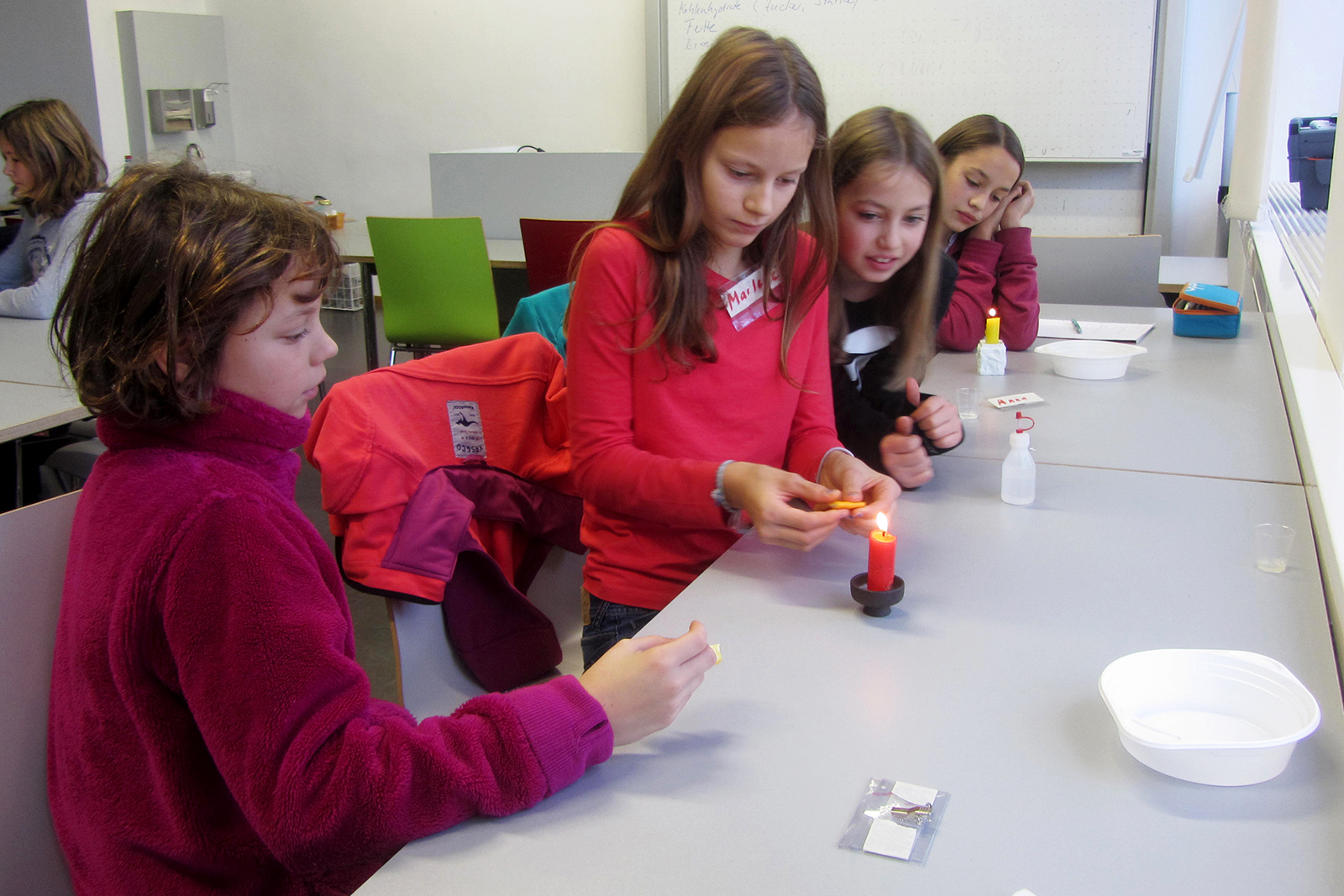 Vier Kinder sitzen um einen Tisch in der Hochschule Coburg und führen ein wissenschaftliches Experiment mit einer brennenden Kerze durch. Ein Kind hält ein Streichholz, während die anderen eifrig zuschauen. Auf dem Tisch in ihrer Nähe sind verschiedene Materialien verstreut, im Hintergrund sind Regale und Stühle zu sehen.