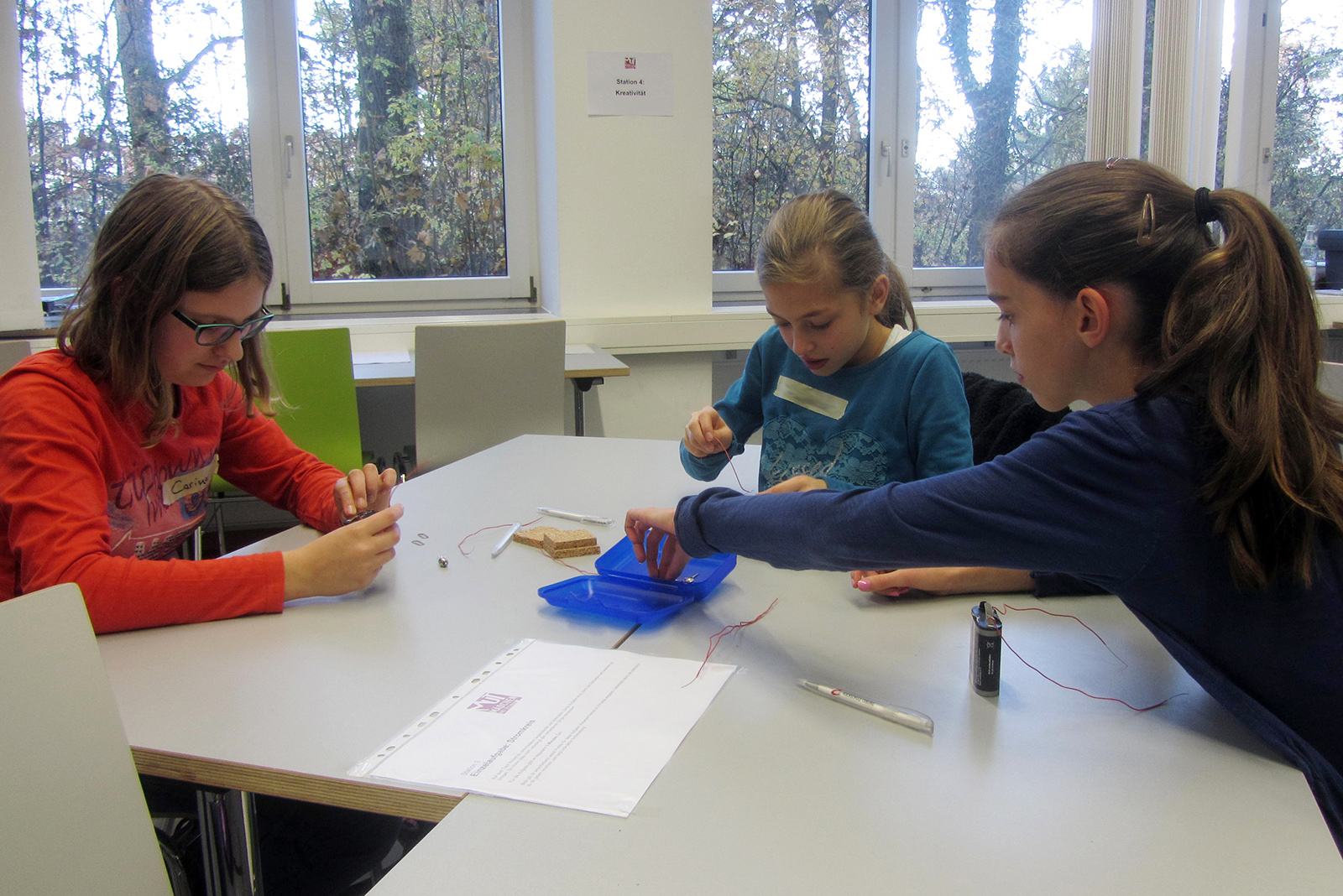 Drei junge Mädchen sitzen an einem Tisch in einem Hörsaal der Hochschule Coburg und arbeiten an einem wissenschaftlichen Projekt mit Batterien, Drähten und einem kleinen Behälter. Sie wirken konzentriert und engagiert bei ihrer Tätigkeit.