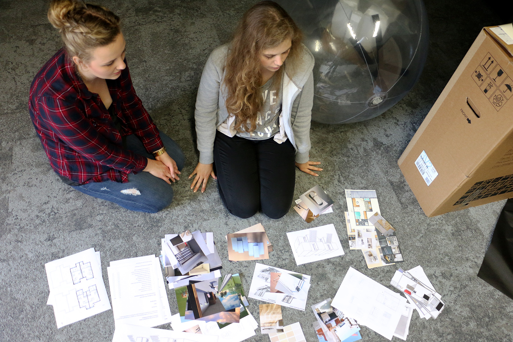 Zwei Personen sitzen auf einem mit Teppich ausgelegten Boden in der Hochschule Coburg, umgeben von verstreuten Papieren und Fotos. Sie arbeiten wahrscheinlich an einem Projekt. Ein großer aufblasbarer Ball und ein Karton in der Nähe deuten auf die kreative Atmosphäre dieses lebhaften akademischen Umfelds hin.