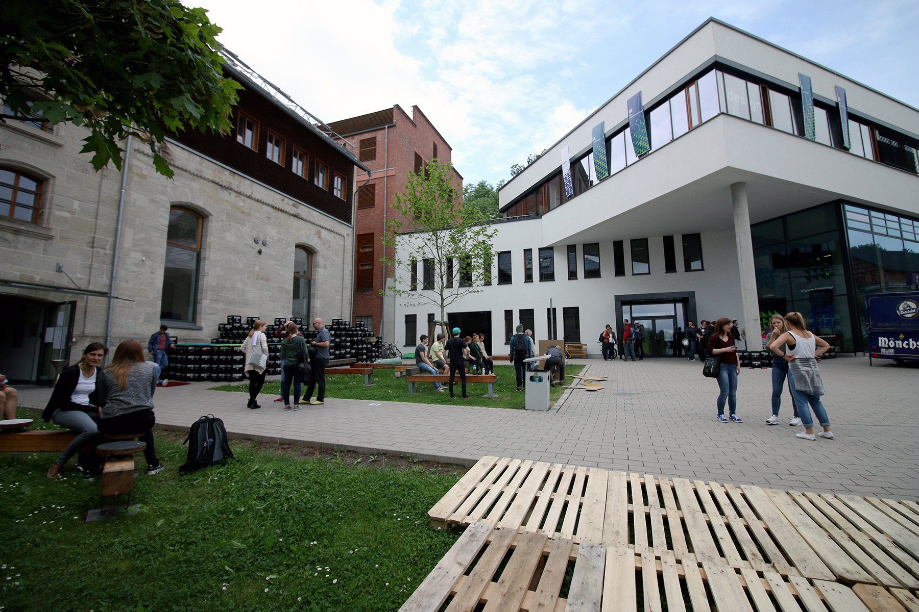 Eine Gruppe von Menschen hat sich vor dem Gebäudekomplex der Hochschule Coburg versammelt, der aus modernen und älteren Gebäuden besteht. Sie sitzen und stehen auf einem gepflasterten Innenhof mit grünem Gras in der Nähe. Holzpaletten und Bäume verleihen der Szenerie unter einem teilweise bewölkten Himmel einen besonderen Charme.