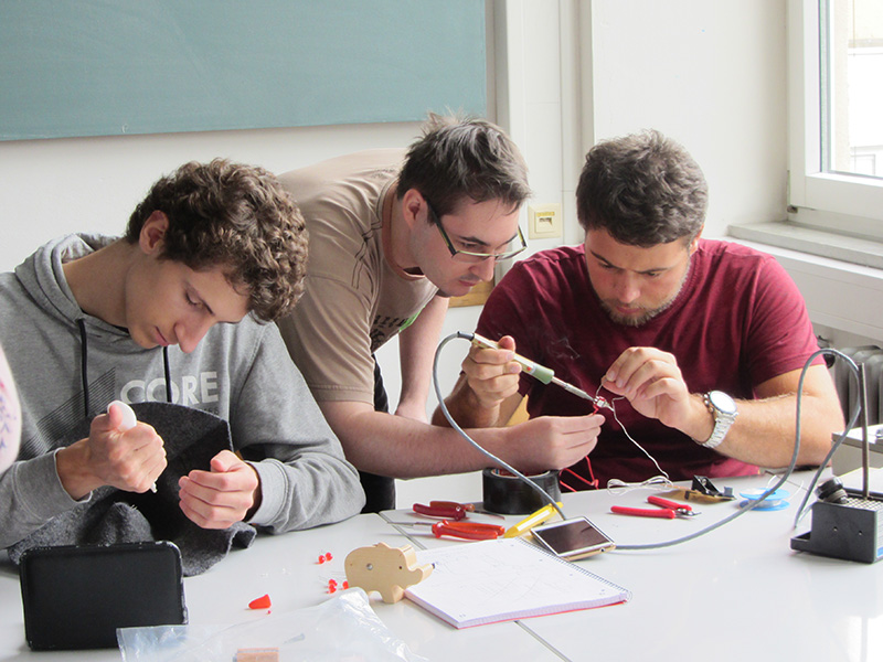 Drei junge Männer arbeiten gemeinsam an einem Tisch in der Hochschule Coburg an einem Elektronikprojekt. In einem hellen Raum mit großen Fenstern löten sie konzentriert Bauteile, die mit Drähten verbunden sind, umgeben von Werkzeug und Materialien.