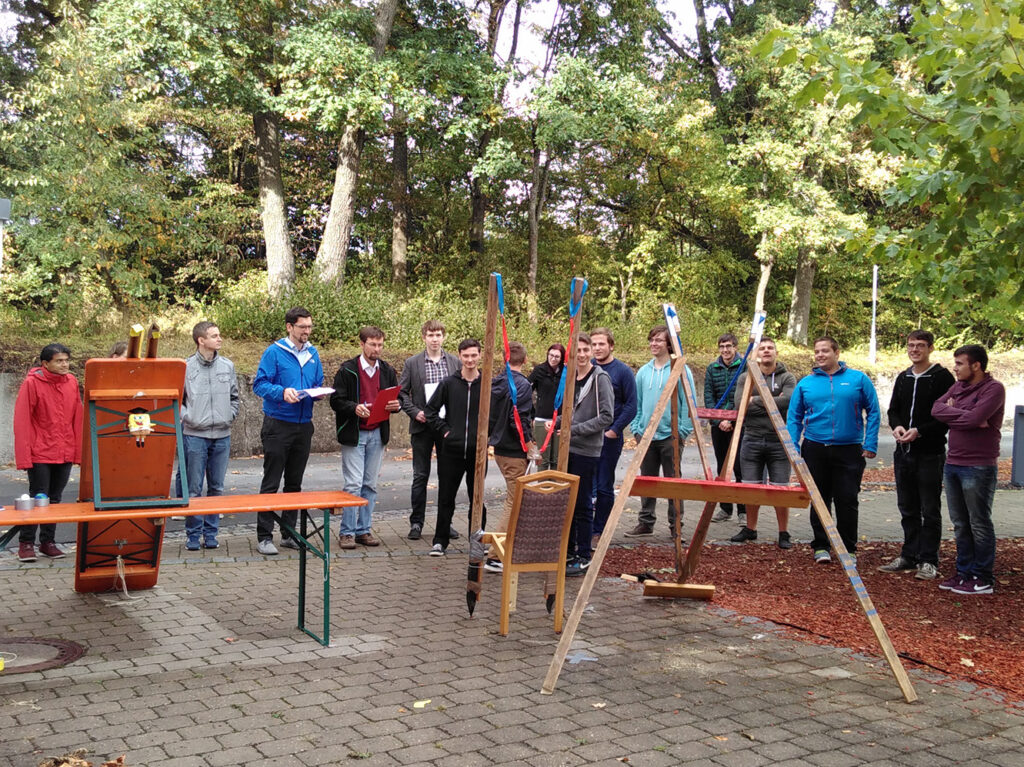 Auf einer gepflasterten Fläche im Freien, umgeben von Bäumen, steht eine Gruppe von Menschen im Halbkreis um provisorische Holzkonstruktionen herum. Einige halten Unterlagen der Hochschule Coburg in den Händen, die Atmosphäre wirkt locker und engagiert.