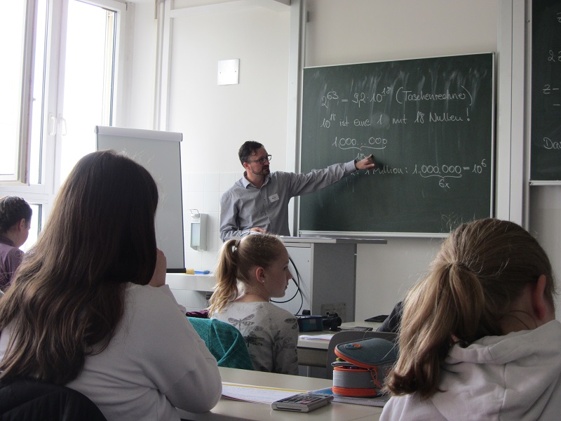 Ein Lehrer der Hochschule Coburg steht vor einem Klassenraum und zeigt auf eine Tafel voller mathematischer Aufgaben. Die Schüler sitzen aufmerksam an ihren Tischen, beobachten und hören zu, während Tageslicht durch ein Fenster auf der linken Seite einfällt.