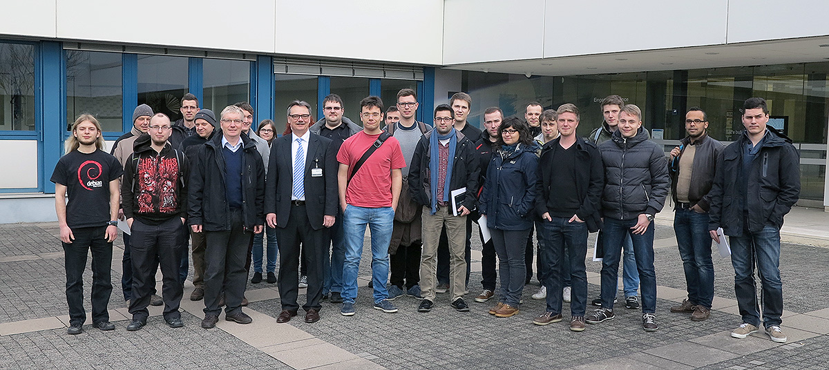 Eine Gruppe von Menschen steht vor einem modernen Gebäude mit großen Fenstern der Hochschule Coburg. Die bunt gemischte Gruppe, bestehend aus Männern und Frauen in Anzügen und Freizeitkleidung, posiert gemeinsam auf einer gepflasterten Fläche.