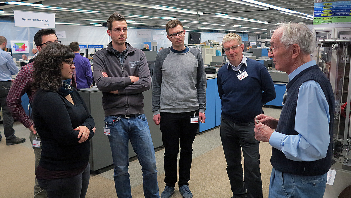 Eine Gruppe von sechs Personen steht im Halbkreis in einer modernen Industrieanlage. Ein älterer Mann in blauer Weste, vermutlich von der Hochschule Coburg, unterhält sich mit fünf aufmerksamen Kollegen in legerer Kleidung. Im Hintergrund sind große Maschinen und Anlagen zu sehen.