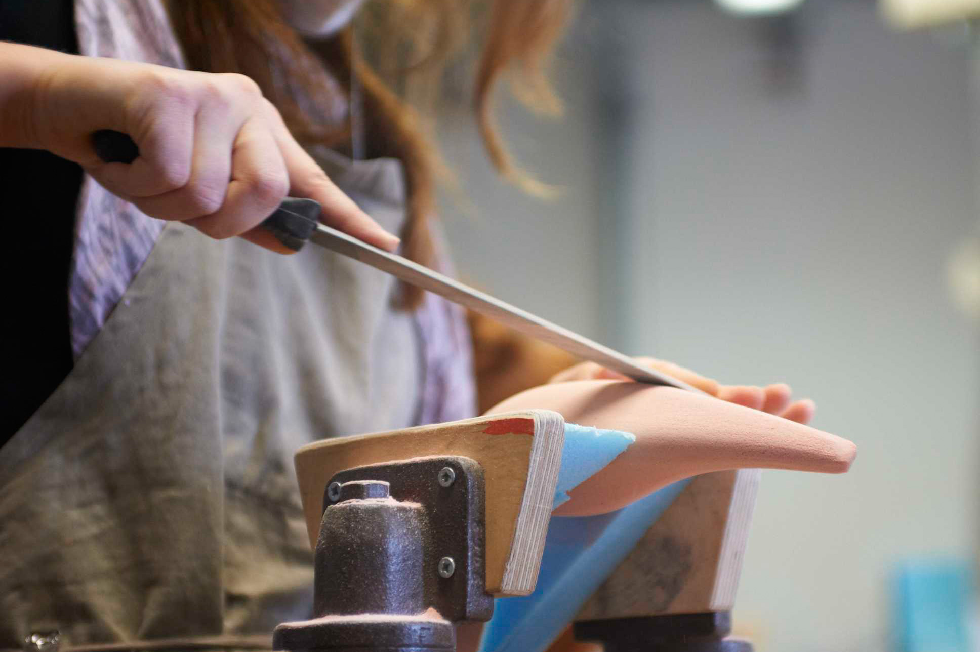 In einer Werkstatt legt eine Person eine Schürze an und formt vorsichtig mit einer Raspel ein Stück Schuhleisten auf der Werkbank. Die handwerkliche Arbeit erinnert an die sorgfältigen Techniken, die an der Hochschule Coburg gelehrt werden.