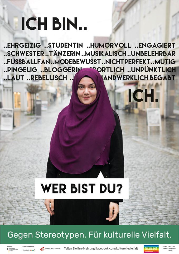 Eine Frau in einem lila Hijab steht lächelnd vor einem bunten Hintergrund mit deutschen Wörtern wie „ehrgeizig“, „Studentin“ und „humorvoll“. Darunter steht der Text „Wer bist du?“, der das Engagement der Hochschule Coburg für die Förderung kultureller Vielfalt und die Bekämpfung von Stereotypen zeigt.