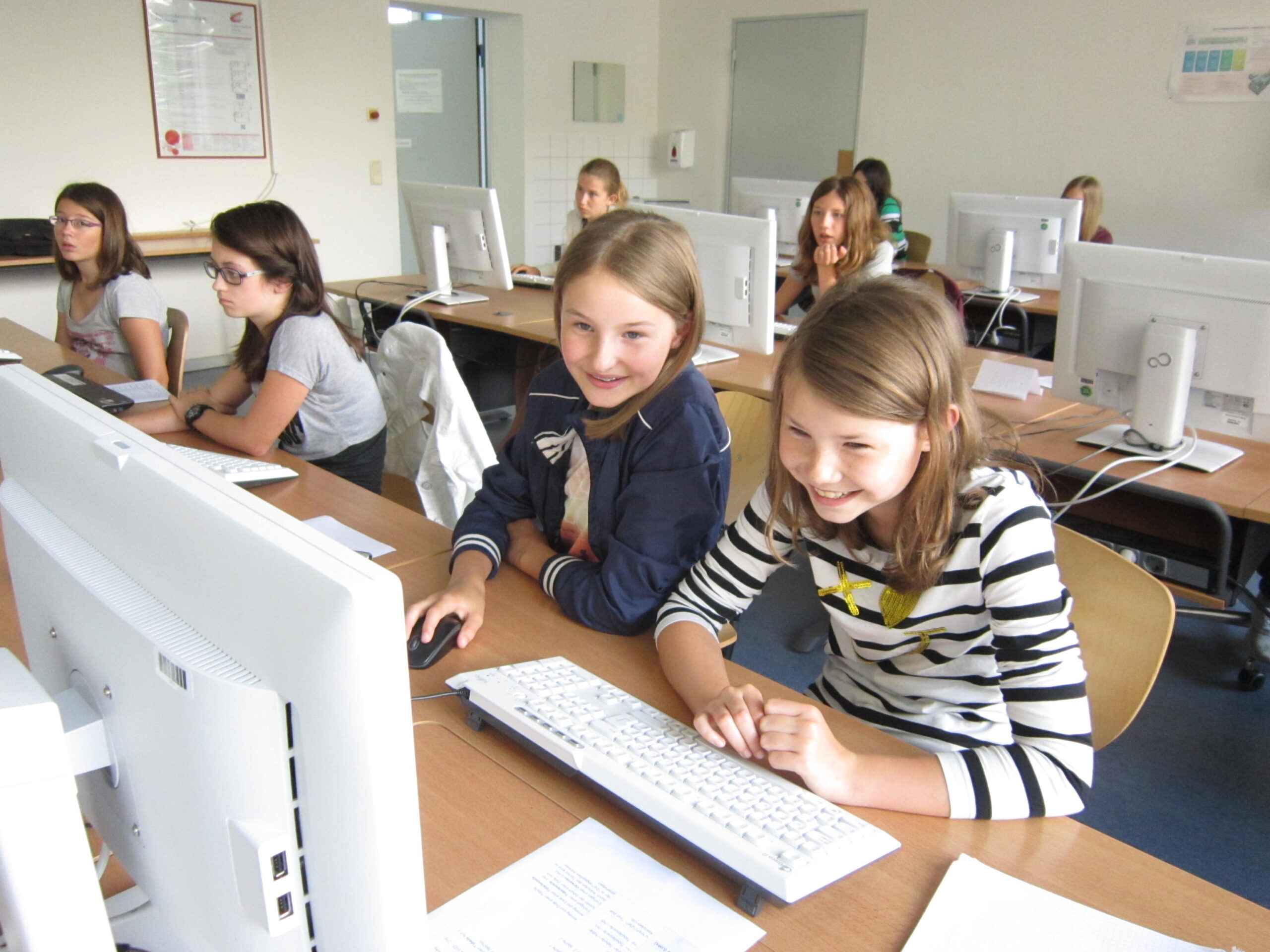 Eine Gruppe junger Studierender sitzt in einem Hörsaal der Hochschule Coburg an Tischen mit Computern. Zwei Mädchen im Vordergrund lächeln und schauen auf einen Monitor. Der Raum, der mit mehreren Computerstationen ausgestattet ist, strahlt eine helle, einladende Atmosphäre aus, die perfekt zum Lernen ist.