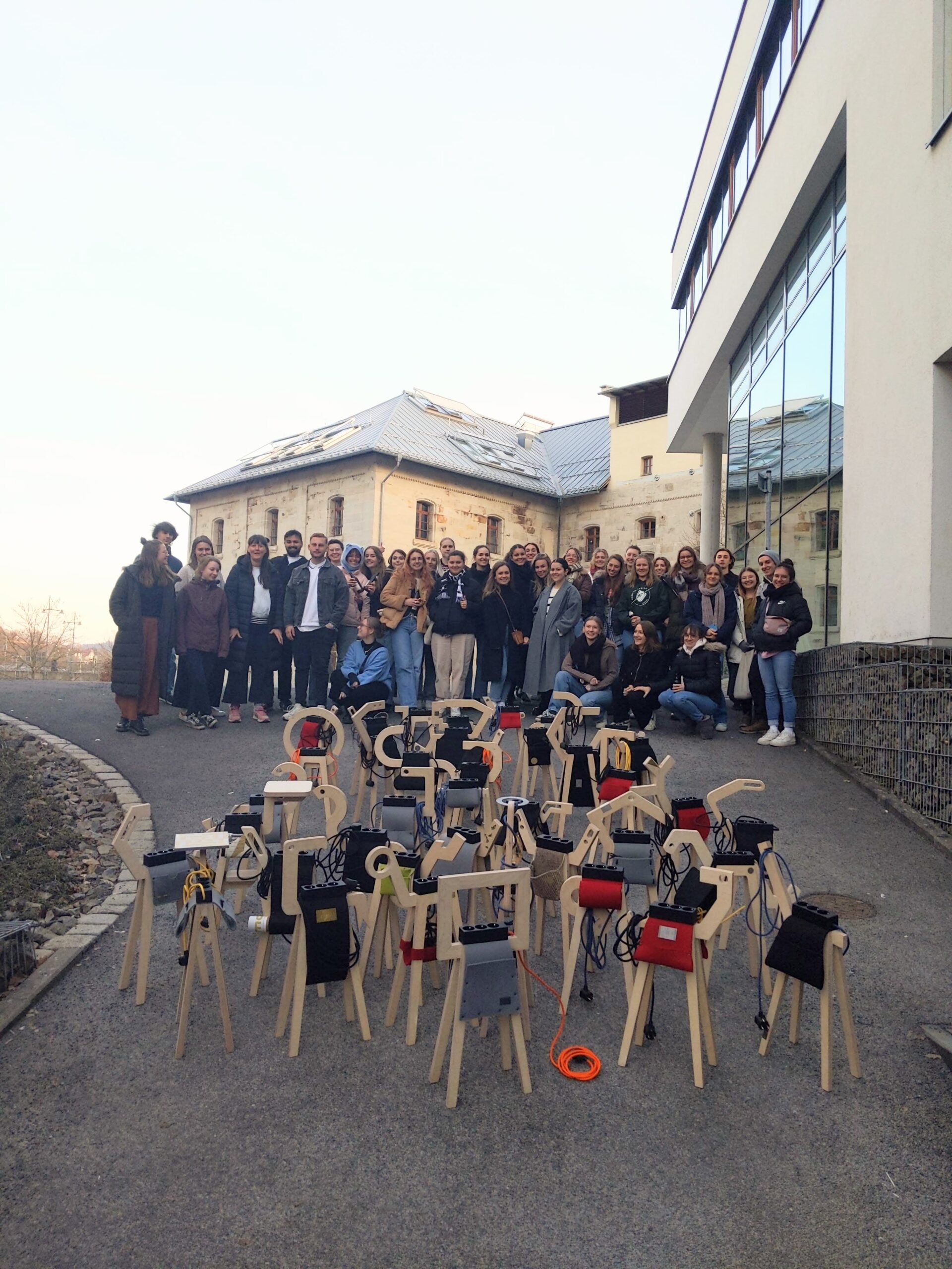 Auf einem Weg neben einem modernen Gebäude der Hochschule Coburg posiert eine große Gruppe von Menschen für ein Foto. Davor sind unter freiem Himmel mehrere kunstvolle Holzskulpturen mit unterschiedlichen Aufsätzen auf dem Boden arrangiert.