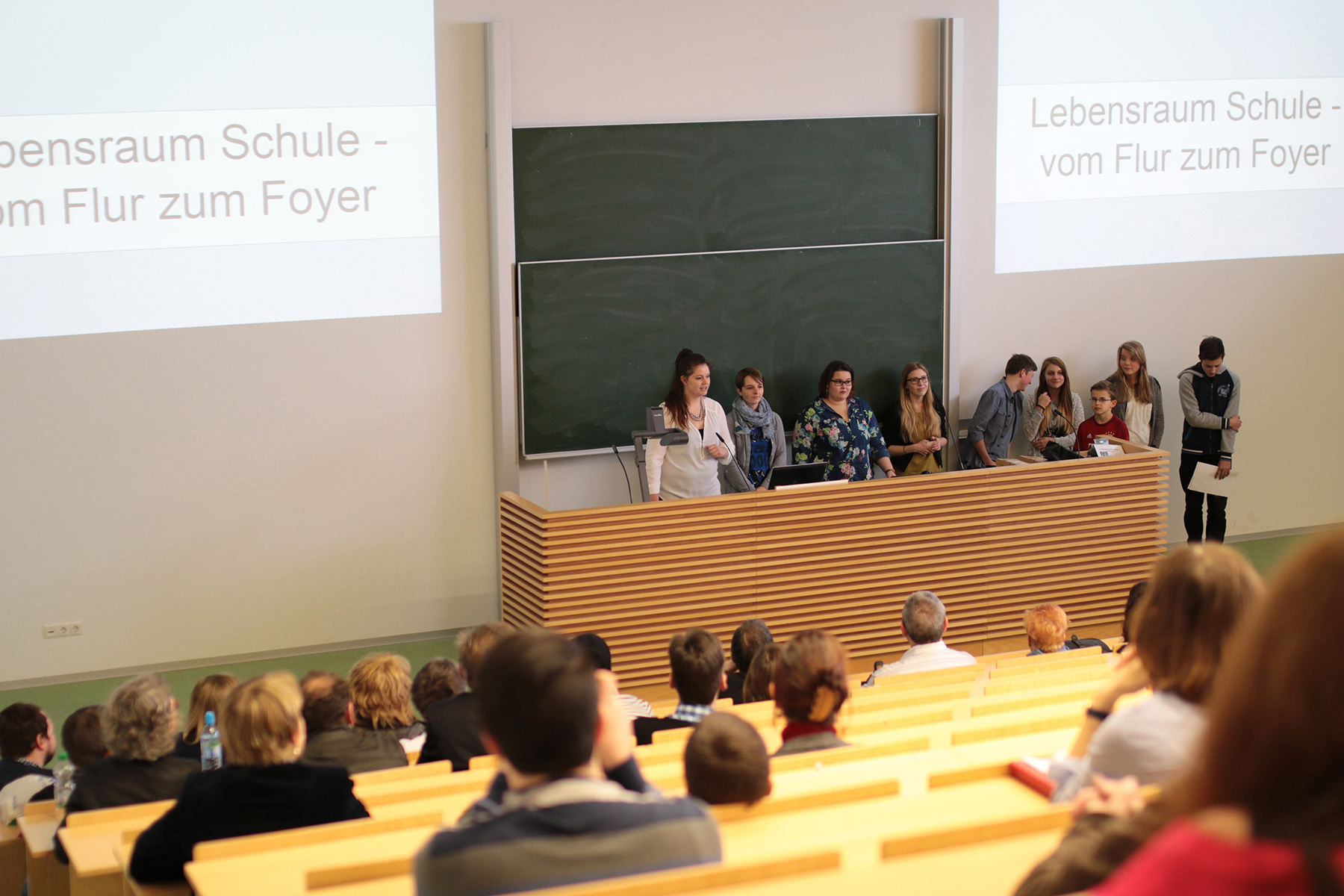 In einem Hörsaal der Hochschule Coburg hält ein Dozent eine Rede an einem Podium vor einer großen Tafel. Die Studenten sitzen in Reihen und hören aufmerksam zu. Auf zwei Projektionsflächen ist ein Text auf Deutsch zu sehen: „Lebensraum Schule – vom Flur zum Foyer“.