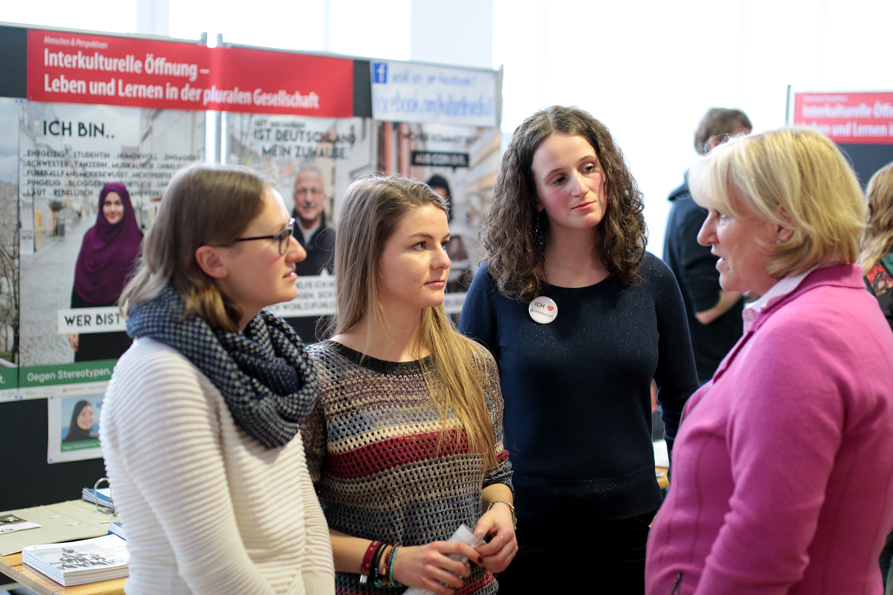 An einem Veranstaltungsstand der Hochschule Coburg unterhalten sich vier Frauen. Dahinter hängen Plakate zum Thema interkultureller Dialog und Lernen in einer vielfältigen Gesellschaft. Die Atmosphäre ist aufmerksam und interaktiv zugleich.