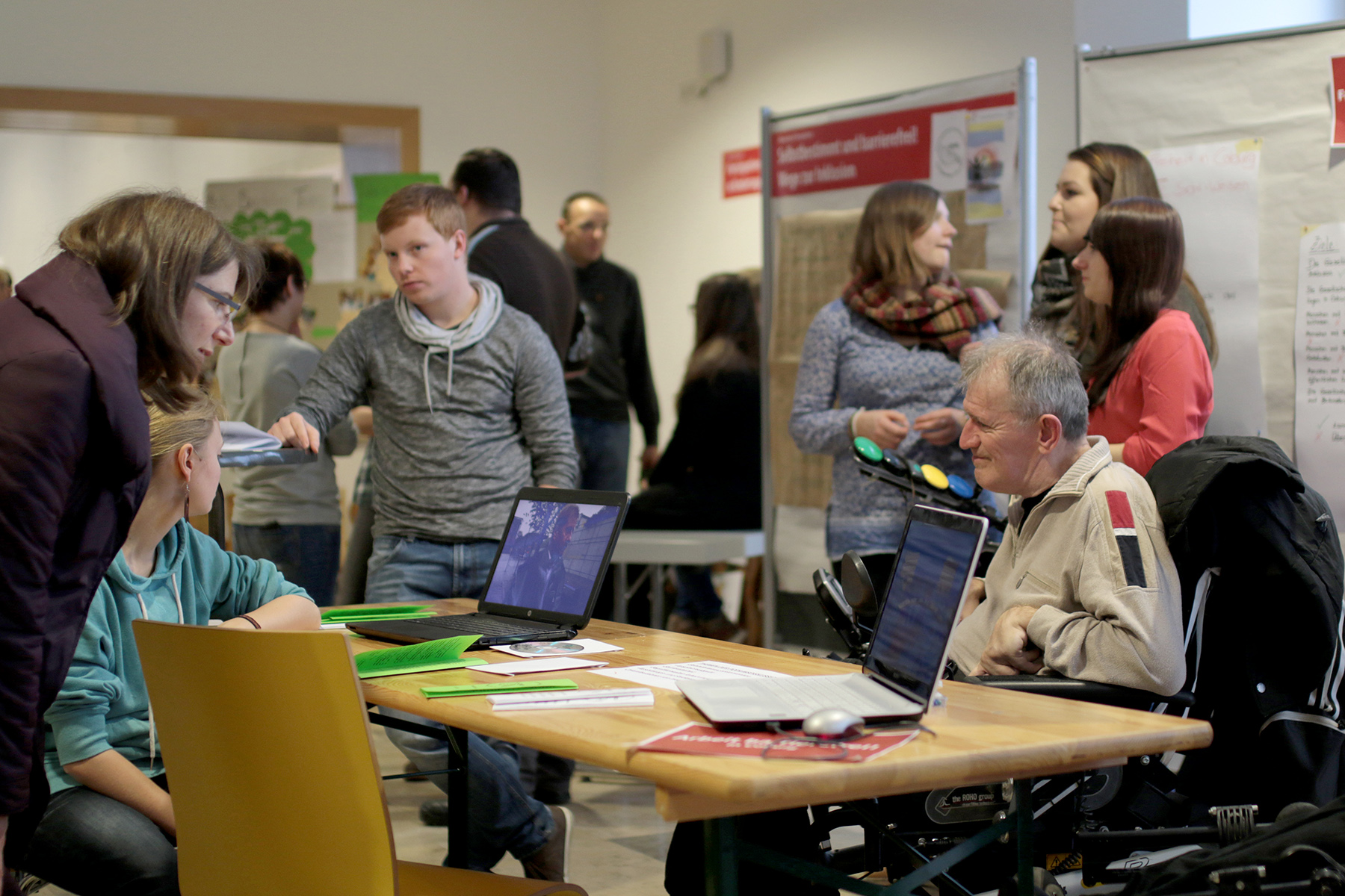 Eine Gruppe von Menschen an der Hochschule Coburg diskutiert an Tischen mit Laptops. Ein Mann im Rollstuhl beobachtet aufmerksam den Bildschirm, während andere in der Nähe stehen und sitzen, interagieren und Materialien lesen. Im Raum herrscht reges Treiben mit verschiedenen Aktivitäten und Notiztafeln.