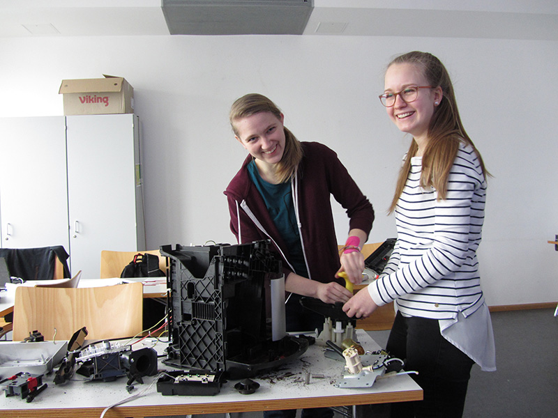 Zwei Personen lächeln und montieren oder reparieren ein Gerät auf einem mit Werkzeugen und Teilen übersäten Tisch. Sie befinden sich in einem gut beleuchteten Raum der Hochschule Coburg mit Stühlen und einem Schrank im Hintergrund und genießen die kollaborative Atmosphäre.