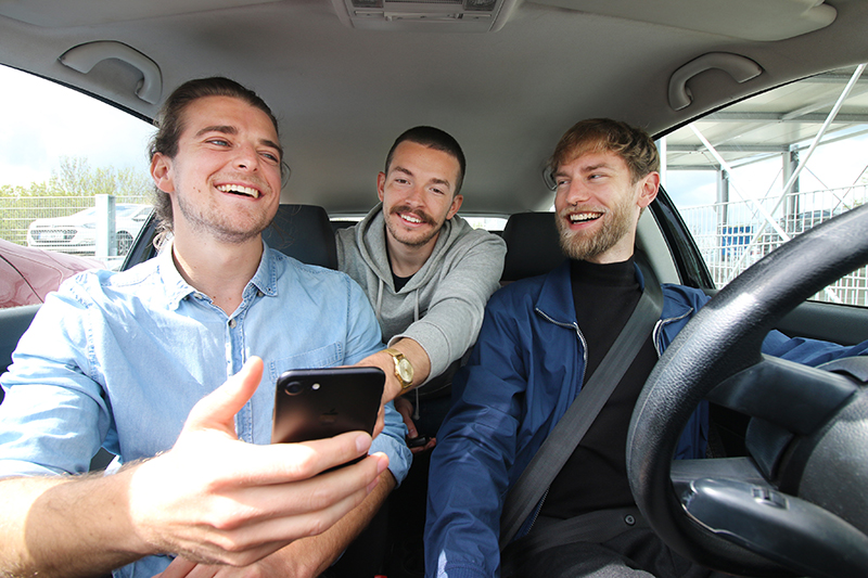 Drei Männer sitzen in einem Auto, lächeln und schauen auf das Smartphone, das der Mann links in der Hand hält, um vielleicht die neuesten Nachrichten der Hochschule Coburg zu checken. Der Mann in der Mitte lehnt sich vom Rücksitz nach vorne, während der Fahrer lächelnd mitmacht.