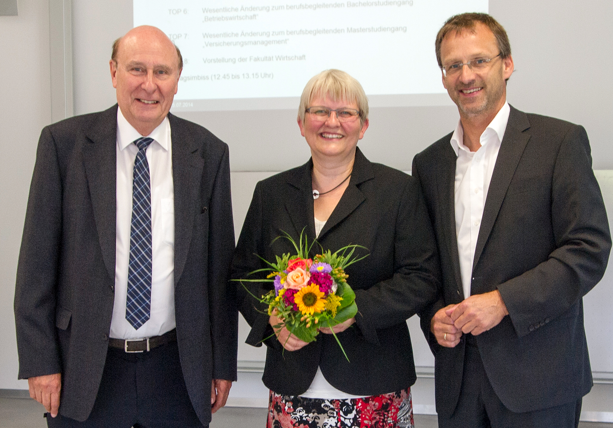 Drei Menschen stehen zusammen und lächeln. Die Person in der Mitte hält einen bunten Blumenstrauß in der Hand. Sie sind in formelle Kleidung gekleidet und stehen vor einem Bildschirm mit der Aufschrift „Hochschule Coburg“.