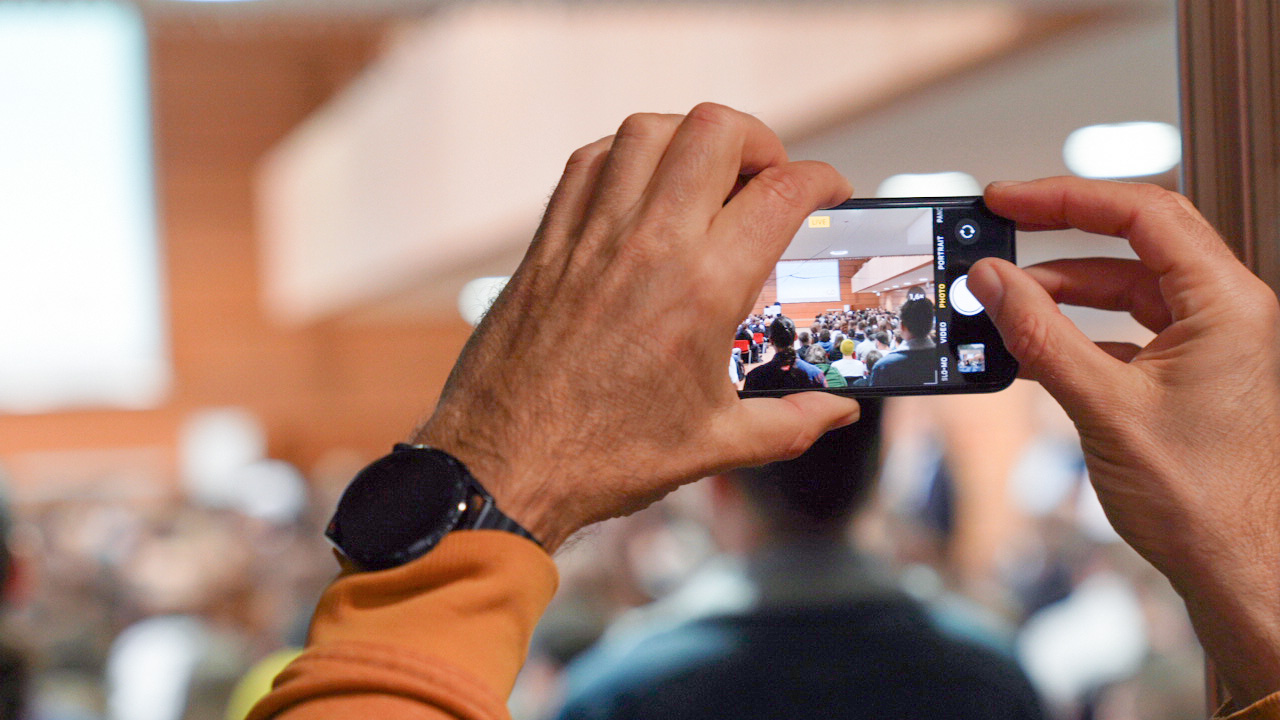 Eine Person in einem gelben Pullover und einer schwarzen Armbanduhr hält ein Smartphone waagerecht und fängt die lebhafte Szene einer Veranstaltung in Innenräumen der Hochschule Coburg ein. Der Smartphone-Bildschirm zeigt die geschäftige Menge im Fokus, mit verschwommenen Teilnehmern und einem Präsentationsbereich im Hintergrund.