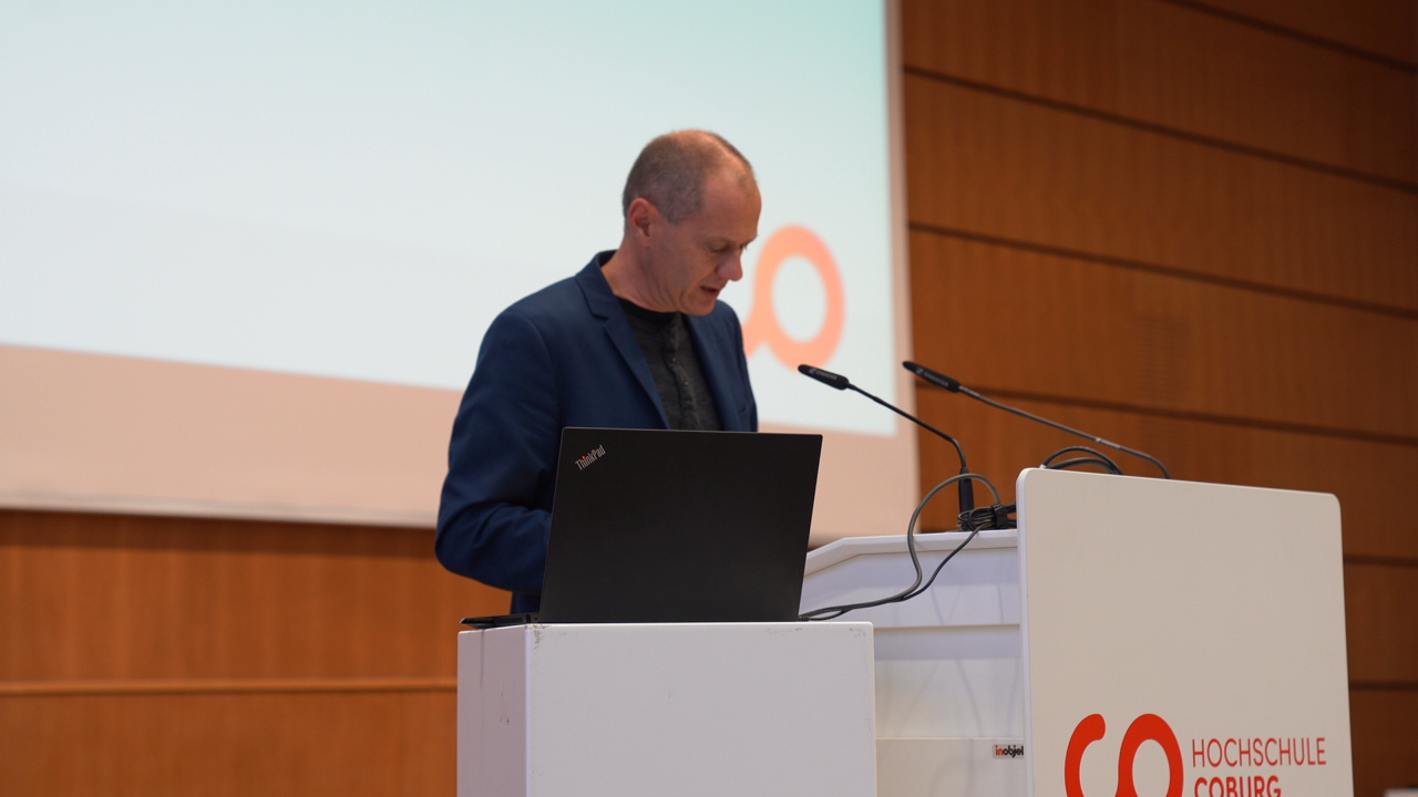 Eine Person steht an einem Podium und hält eine Rede in einem Hörsaal. Mit Mikrofonen und einem Laptop, auf dem prominent „Hochschule Coburg“ steht, strahlt die Umgebung Formalität aus. Ein großer Bildschirm im Hintergrund unterstreicht die Wichtigkeit der Veranstaltung.