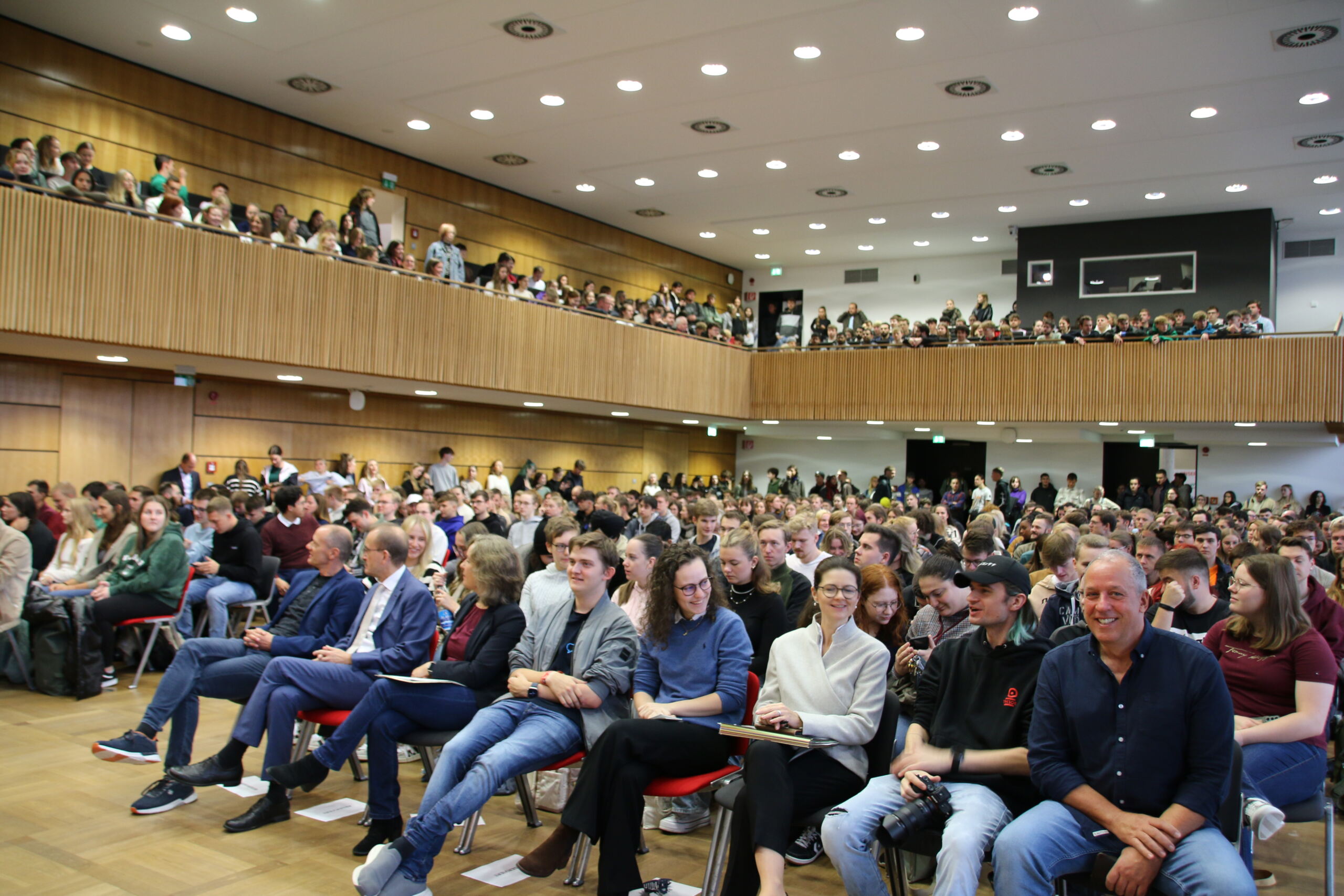 Eine große Gruppe von Menschen sitzt für eine Veranstaltung im geräumigen Hörsaal der Hochschule Coburg. Das Publikum, das aus verschiedenen Altersgruppen besteht, füllt sowohl den Balkon als auch den unteren Sitzbereich. Der Raum verfügt über Holzwände und helles Licht, was eine einladende akademische Atmosphäre schafft.