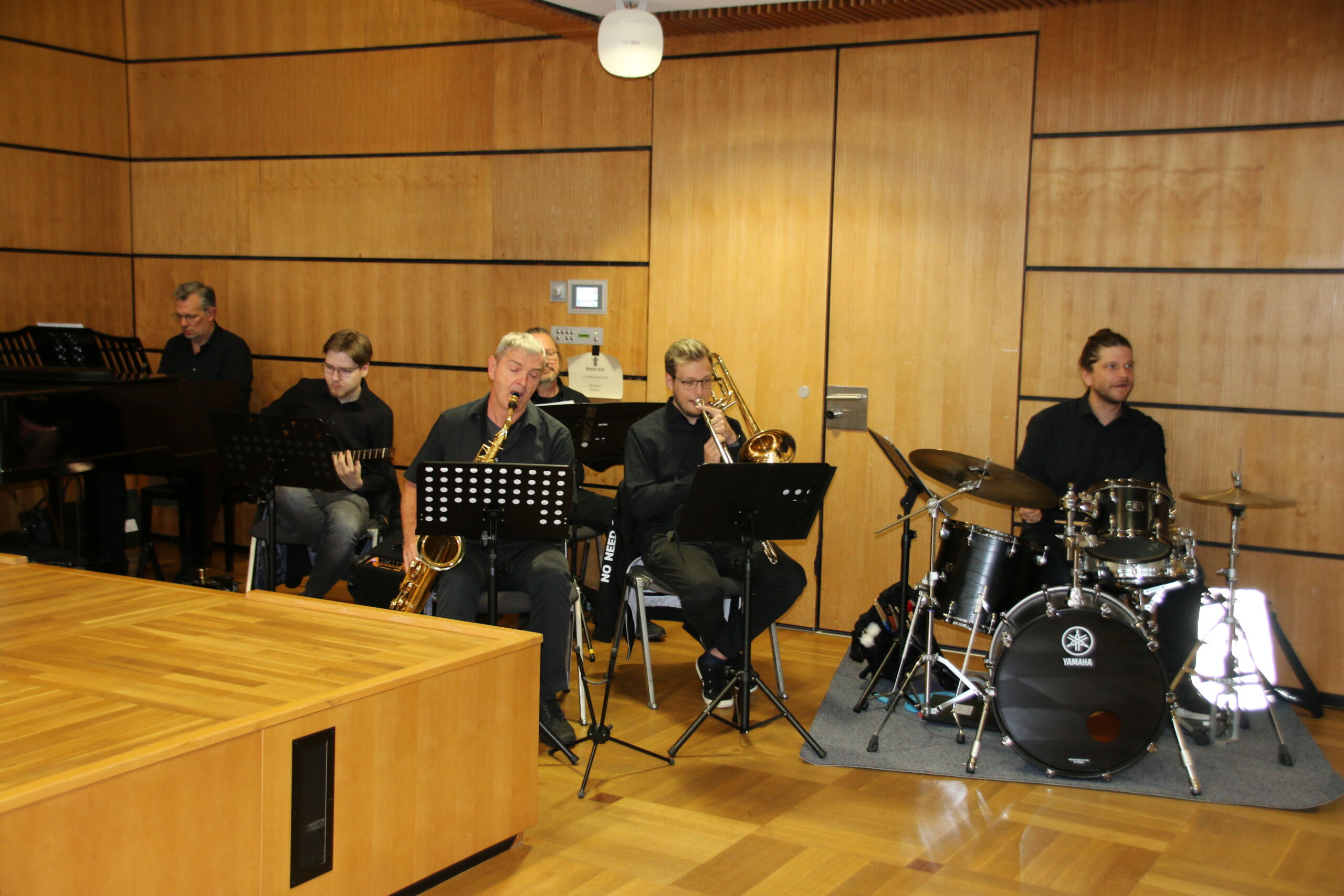 Eine Band der Hochschule Coburg tritt im Innenbereich auf. Die Gruppe besteht aus einem Pianisten, einem Saxophonisten, einem Posaunisten, einem Gitarristen und einem Schlagzeuger. Sie sind dunkel gekleidet und sitzen neben einer Holzwand mit Notenständern, auf denen ihre Notenblätter liegen.