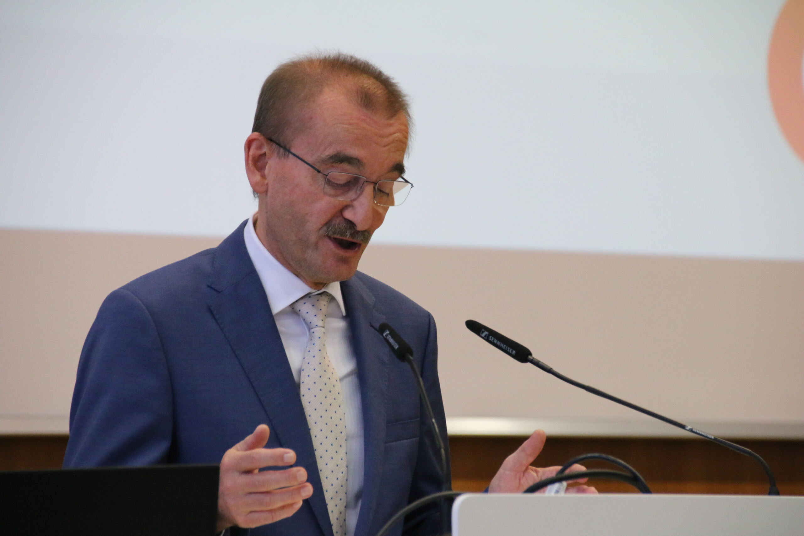 Eine Person mit Brille und blauem Anzug spricht leidenschaftlich an einem Podium mit Mikrofon. Sie gestikuliert mit den Händen und im Hintergrund ist ein Bildschirm mit der Aufschrift „Hochschule Coburg“ zu sehen.