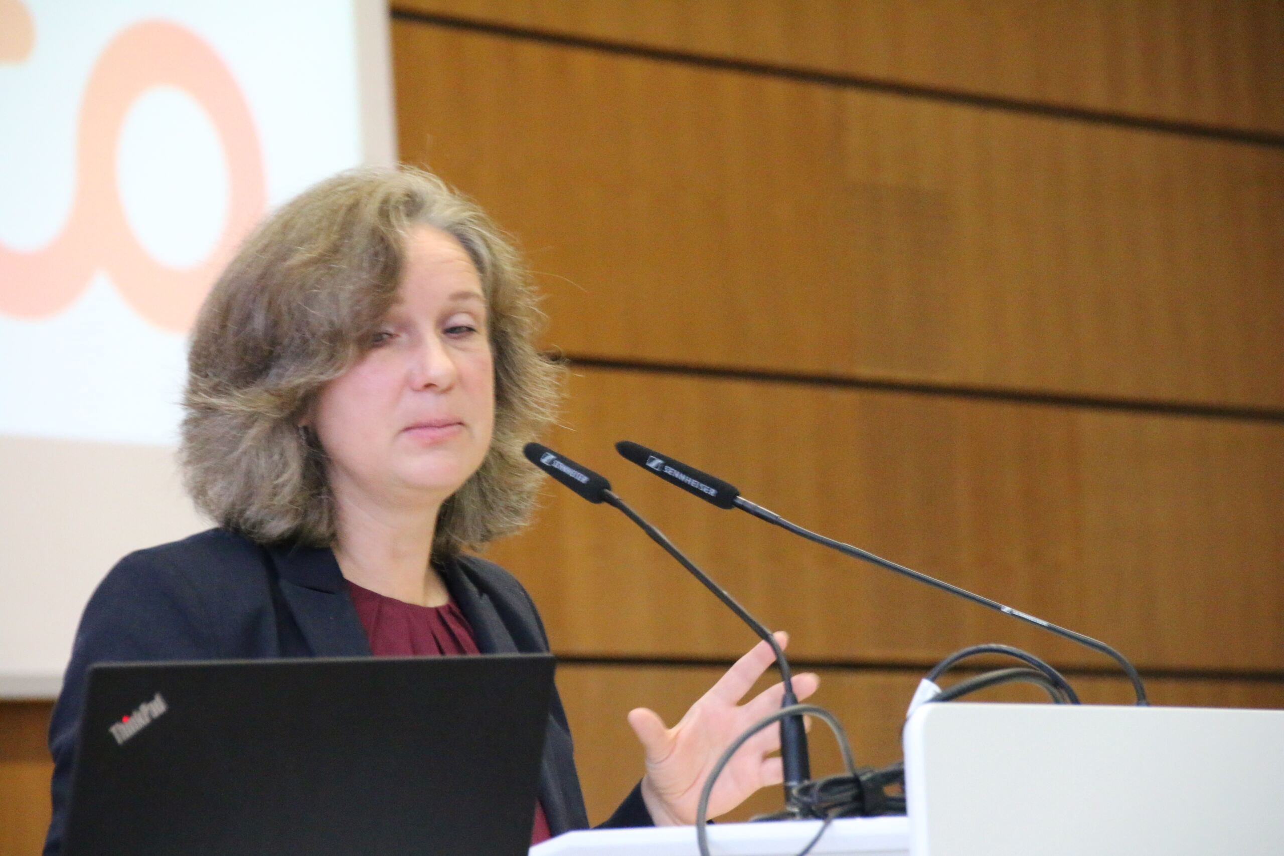 Eine Person mit schulterlangem Haar spricht an einem Podium mit Mikrofonen, flankiert von einem Laptop. Sie befinden sich in einem Raum mit Holzwänden, auf der Projektionsfläche ist ein Teil des Logos der Hochschule Coburg zu sehen.