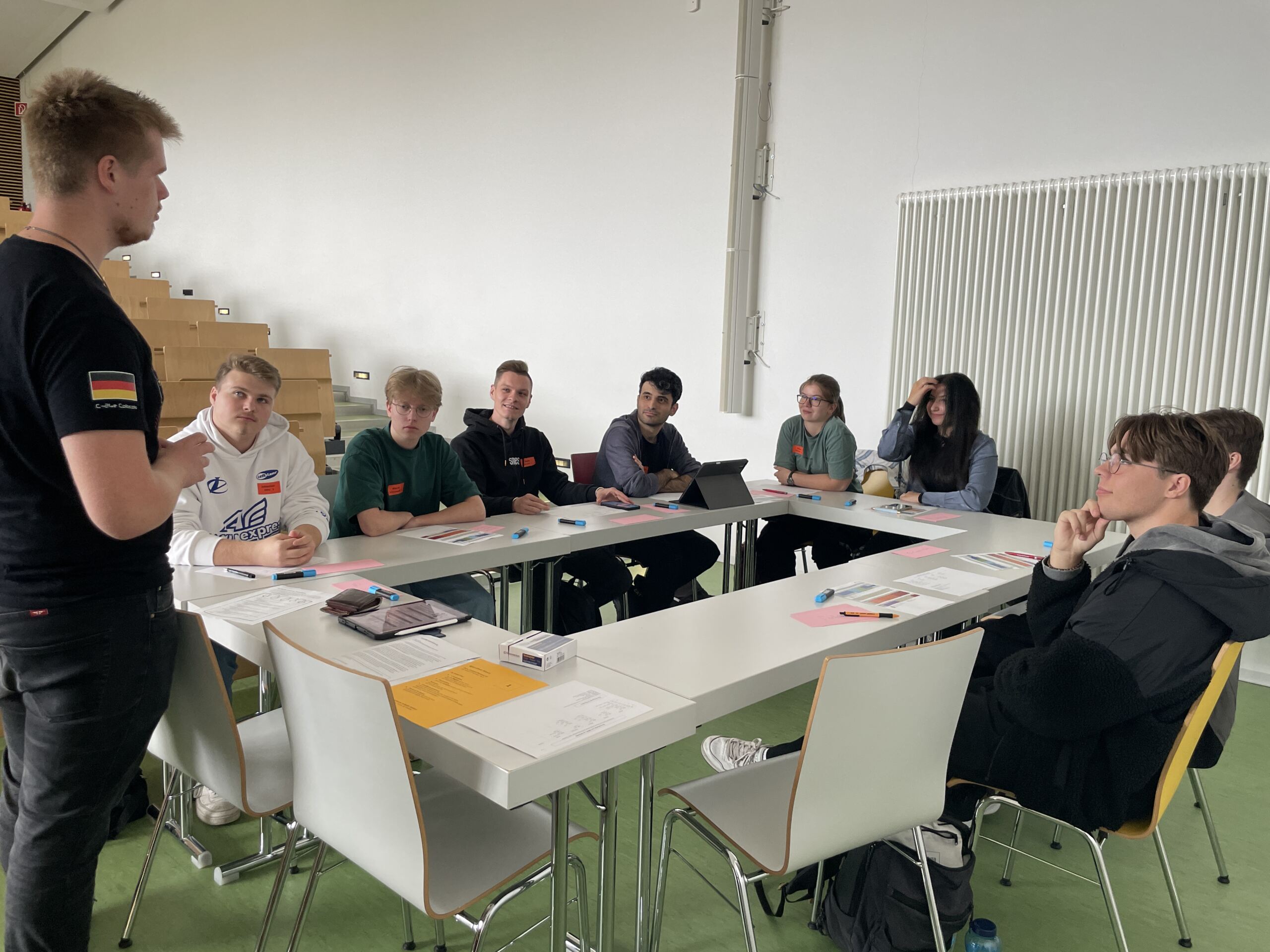 Eine Gruppe von Menschen sitzt in einem Klassenzimmer um einen Tisch herum und führt eine lebhafte Diskussion unter der Leitung eines Redners. Papier und Stifte liegen verstreut herum, während sie sich voller Enthusiasmus auf die Erstsemestertage vorbereiten und bereit sind, gemeinsam mit Spaß und Kameradschaft in dieses neue Kapitel zu starten.