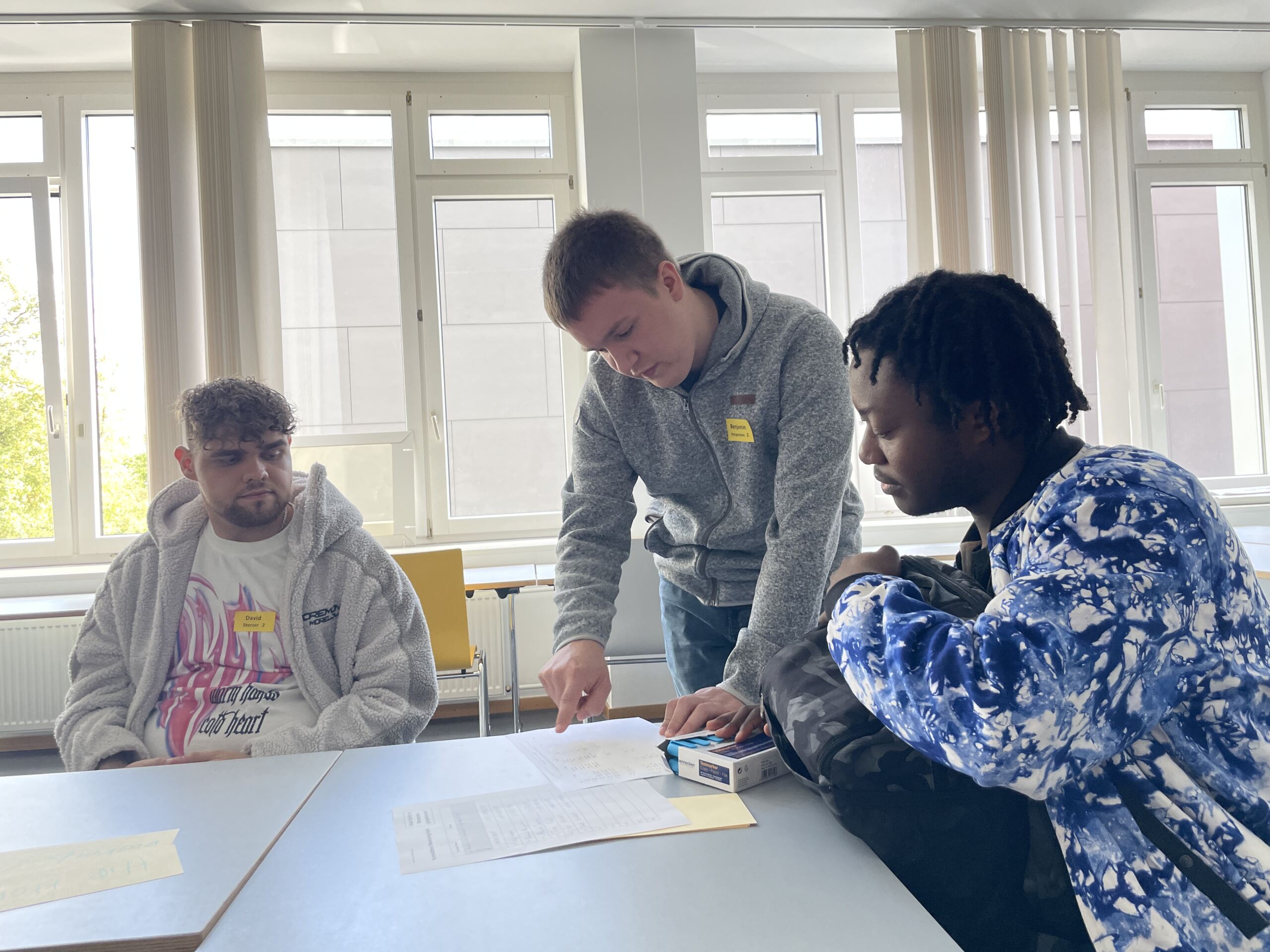 In einem hell erleuchteten Raum diskutieren drei Personen lebhaft und zeigen auf ein Papier auf dem Tisch. Die Atmosphäre erinnert an die Kameradschaft der Erstsemestertage, während sie an großen Fenstern zusammenarbeiten und darauf brennen, gemeinsam ihre Reise zu beginnen.