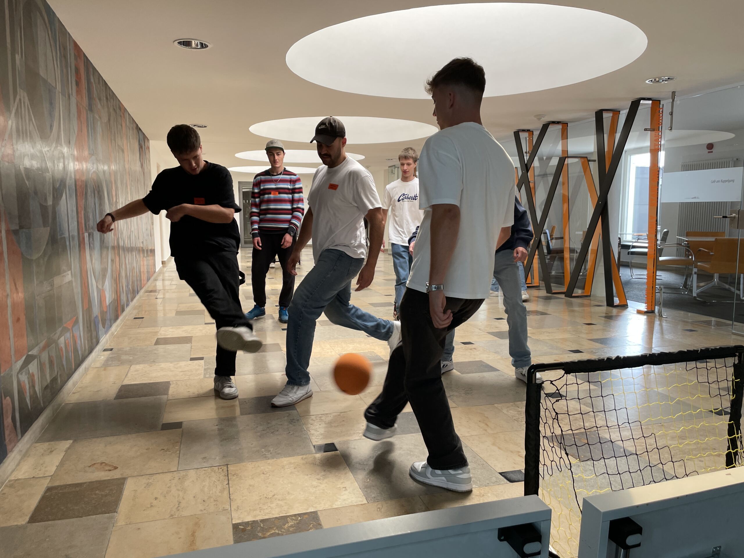 Eine Gruppe von sieben Studierenden spielt während der Erstsemestertage in einem geräumigen Raum mit Marmorboden ein lockeres Hallenfußballspiel. Sie konzentrieren sich auf den Ball und zielen auf kleine Tore an beiden Enden. Die Wände sind mit abstrakter Kunst geschmückt und natürliches Licht trägt zur fröhlichen Atmosphäre bei.