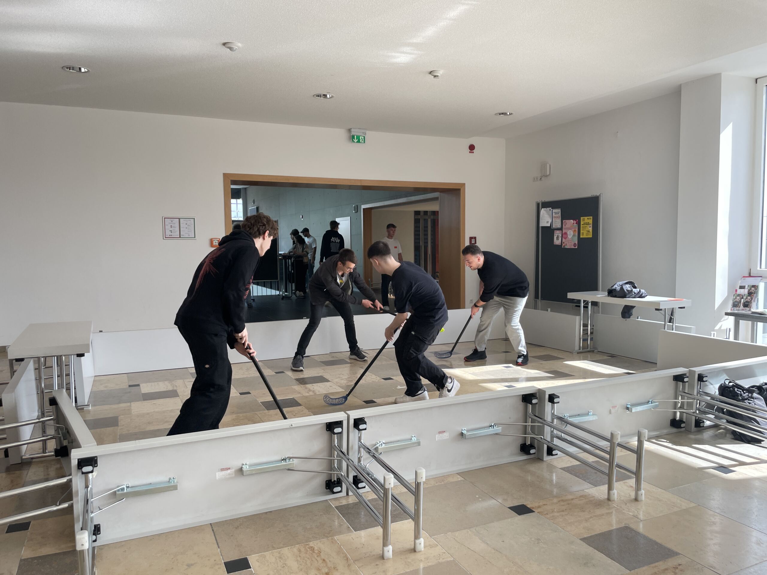 Vier Personen spielen in einem hellen Raum Tischhockey und haben gemeinsam Spaß. Das Spiel wird auf einem rechteckigen Tisch mit verstellbaren Seiten aufgebaut. Sonnenlicht fällt durch ein großes Fenster und erhellt den Raum, während ein paar andere aus der Ferne zuschauen und den Spaß dieses gemeinschaftlichen Moments teilen.