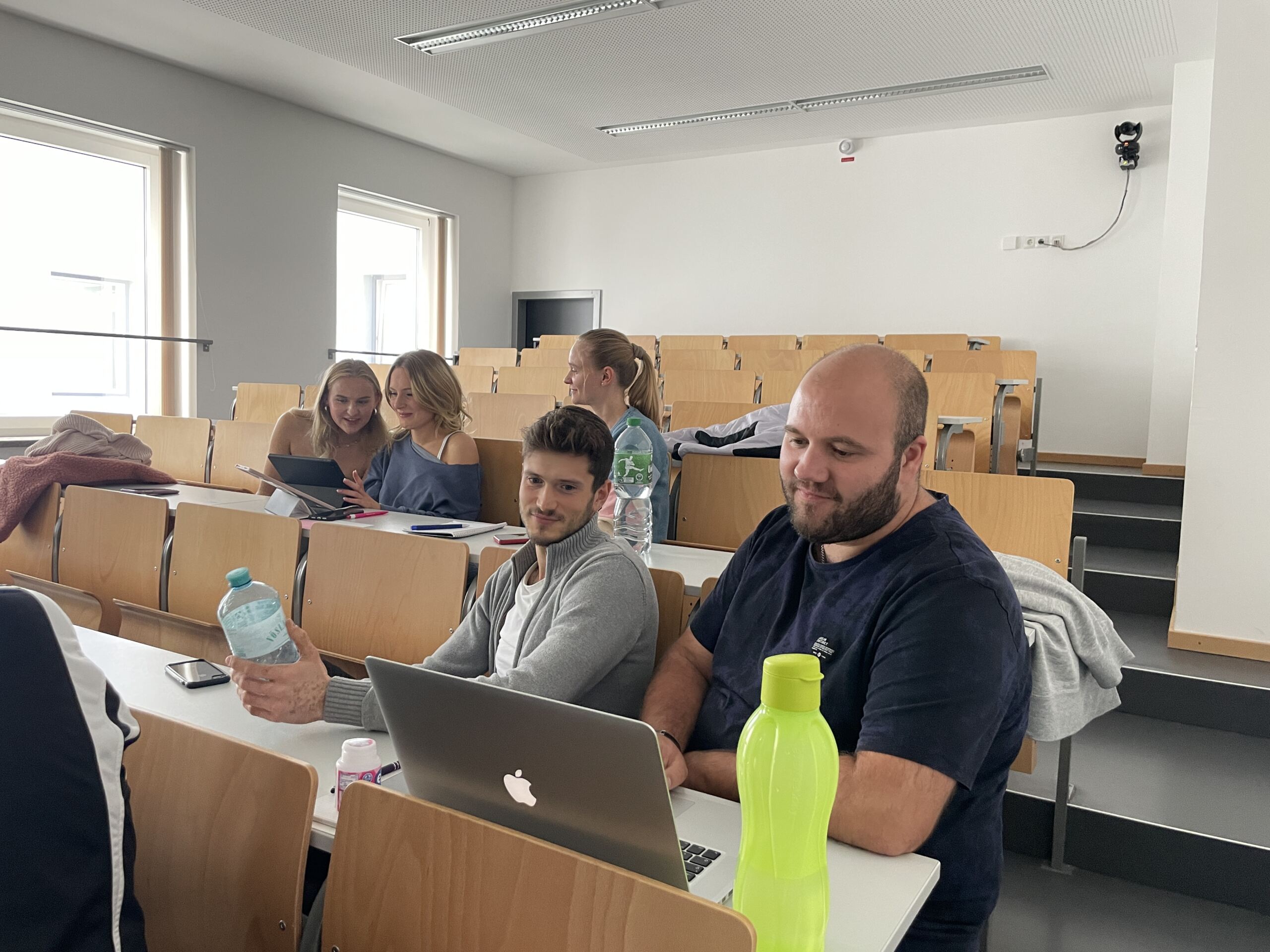 Im Vorlesungssaal herrscht reges Treiben. Mehrere Studierende sitzen in Reihen von Holztischen. Einige nutzen Laptops und Tablets, während andere begeistert über ihre Erfahrungen bei den Erstsemestertagen sprechen. Helle Fenster erhellen den Raum und auf dem Tisch im Vordergrund steht eine große grüne Wasserflasche.