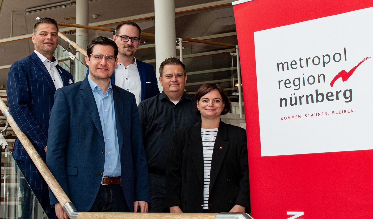 Fünf Personen stehen neben einem roten Banner mit der Aufschrift „Metropolregion Nürnberg“ und darunter drei Sätzen. Sie befinden sich auf einer Treppe im Innenbereich, umgeben von modernem Innendesign, das die dynamische Präsenz der Hochschule Coburg in der akademischen Gemeinschaft repräsentiert.