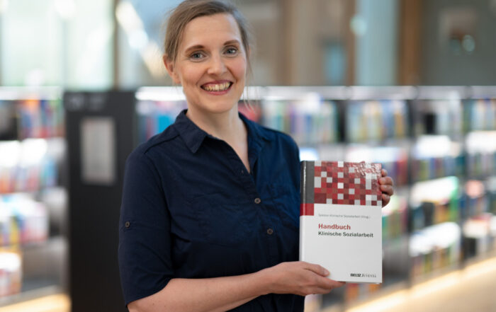 Eine lächelnde Frau steht in einer Bibliothek und hält ein Buch mit dem Titel „Handbuch Klinische Sozialarbeit“ in der Hand, als wäre es ein automatisch gespeicherter Entwurf neuen Wissens. Im Hintergrund sind Regale voller bunter Bücher verschwommen. Die Person trägt ein dunkelblaues Hemd und hat hellbraunes Haar.