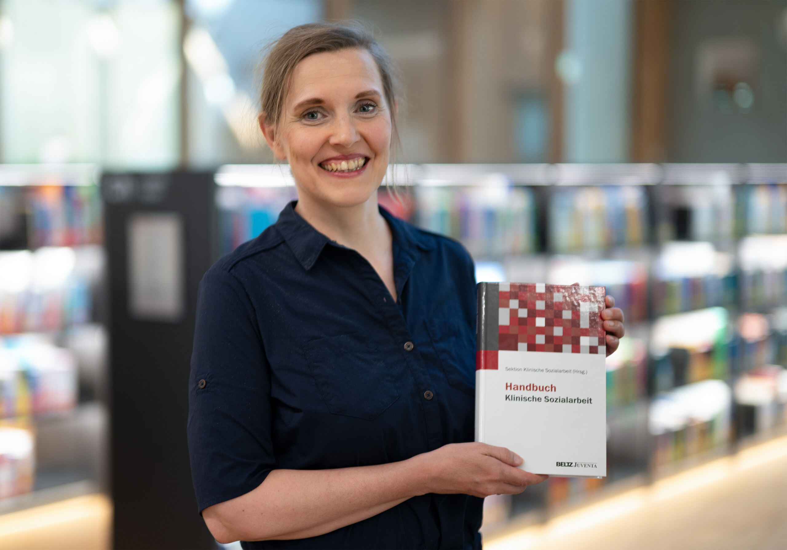 Eine lächelnde Frau steht in einer Bibliothek und hält ein Buch mit dem Titel „Handbuch Klinische Sozialarbeit“ in der Hand, als wäre es ein automatisch gespeicherter Entwurf neuen Wissens. Im Hintergrund sind Regale voller bunter Bücher verschwommen. Die Person trägt ein dunkelblaues Hemd und hat hellbraunes Haar.