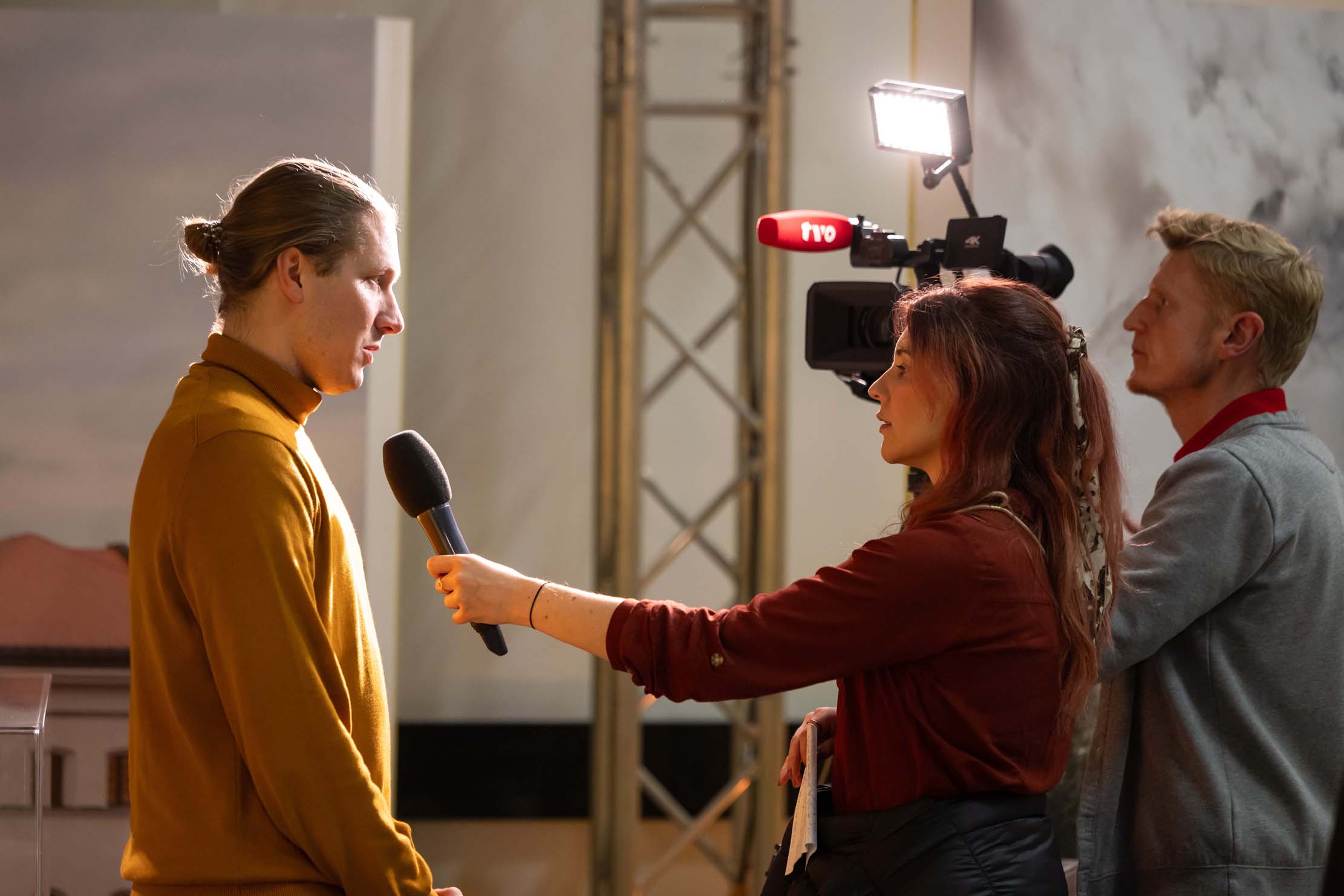 Eine Person mit langen Haaren wird von einer Frau mit Mikrofon interviewt, die die Hochschule Coburg vertritt. Ein Kameramann hält den Moment mit professioneller Videoausrüstung fest, die aus einer Kamera und einem Licht besteht. Die Szene scheint sich in einem Innenbereich vor einem schlichten Hintergrund zu befinden.