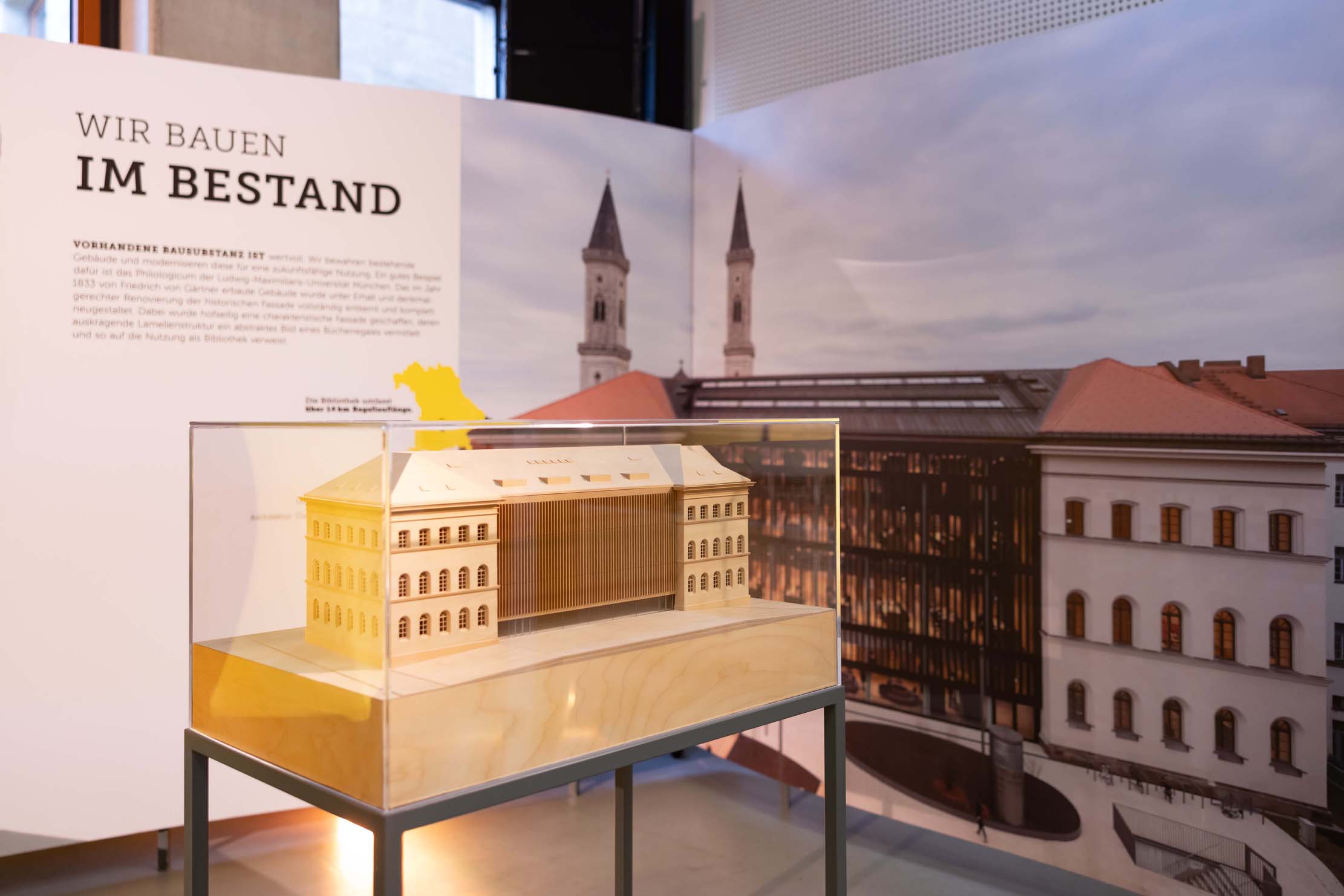 Ein hölzernes Architekturmodell in einer Glasvitrine wird vor einem Hintergrund mit dem deutschen Text „WIR BAUEN IM BESTAND“ ausgestellt. Die Szene würdigt das Ethos der Hochschule Coburg und zeigt eine Gebäudeaußenseite, die zwei Türme miteinander verbindet – eine harmonische Mischung aus moderner und traditioneller Architektur.