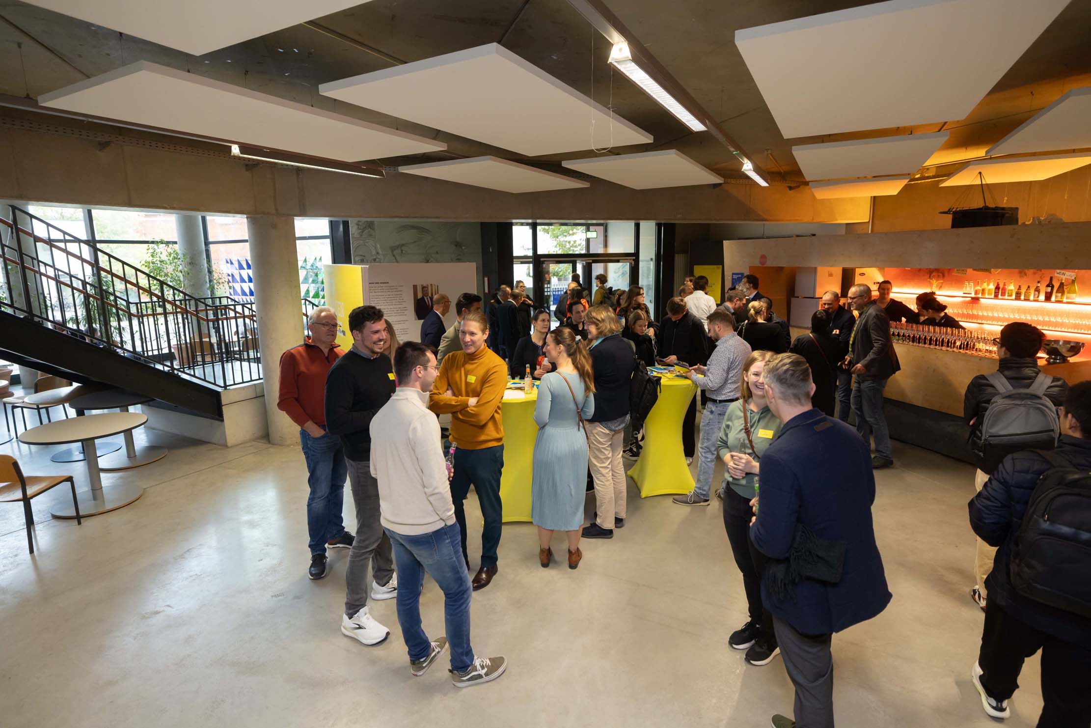 Eine Gruppe von Menschen trifft sich in einem modernen Innenraum der Hochschule Coburg und trifft sich in der Nähe einer Bar. Einige stehen an Tischen, unterhalten sich und halten Getränke in der Hand. Der Raum verfügt über große Fenster, zeitgenössische Möbel und eine Treppe im Hintergrund.