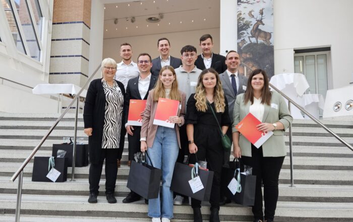 Eine Gruppe von zehn Personen, acht junge Erwachsene und zwei ältere Erwachsene, steht auf einer Treppe der Hochschule Coburg und hält Zertifikate und Geschenktüten in den Händen. Sie befinden sich in einem modernen Gebäude mit einem großen Gemälde und Tageslicht, das durch die Fenster darüber strömt.