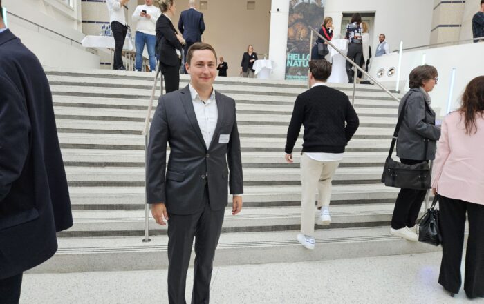 Ein Mann im Anzug steht am Fuß einer breiten Treppe im Innenbereich der Hochschule Coburg. Mehrere Personen sind auf der Treppe und im Hintergrund und unterhalten sich. Die Umgebung wirkt formell oder professionell und spiegelt eine Atmosphäre akademischer Exzellenz wider.